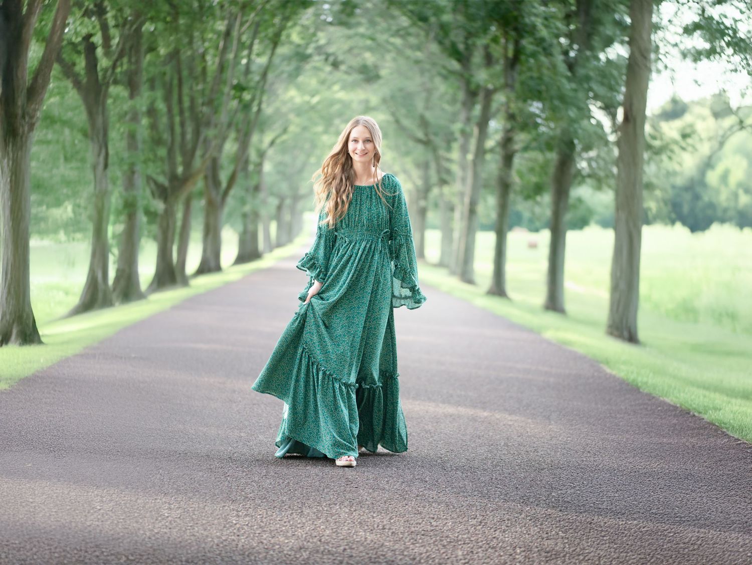 a pretty blonde senior student in a long green dress strolls toward the camera down a tree-lined street surrounded by fields