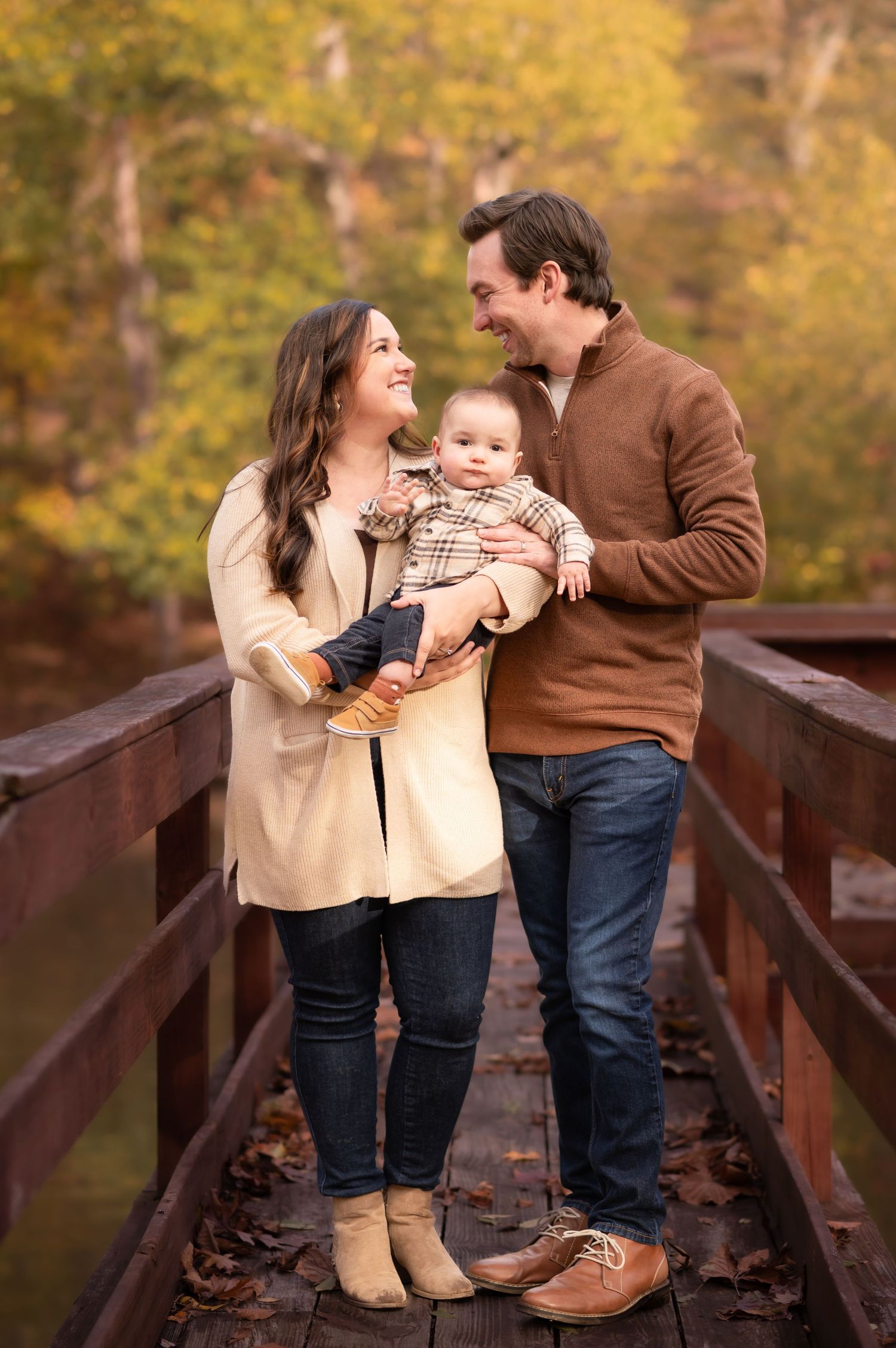 a cute little family of three pose together on a wooden bridge with fall leaves behind them in Morgantown, WV