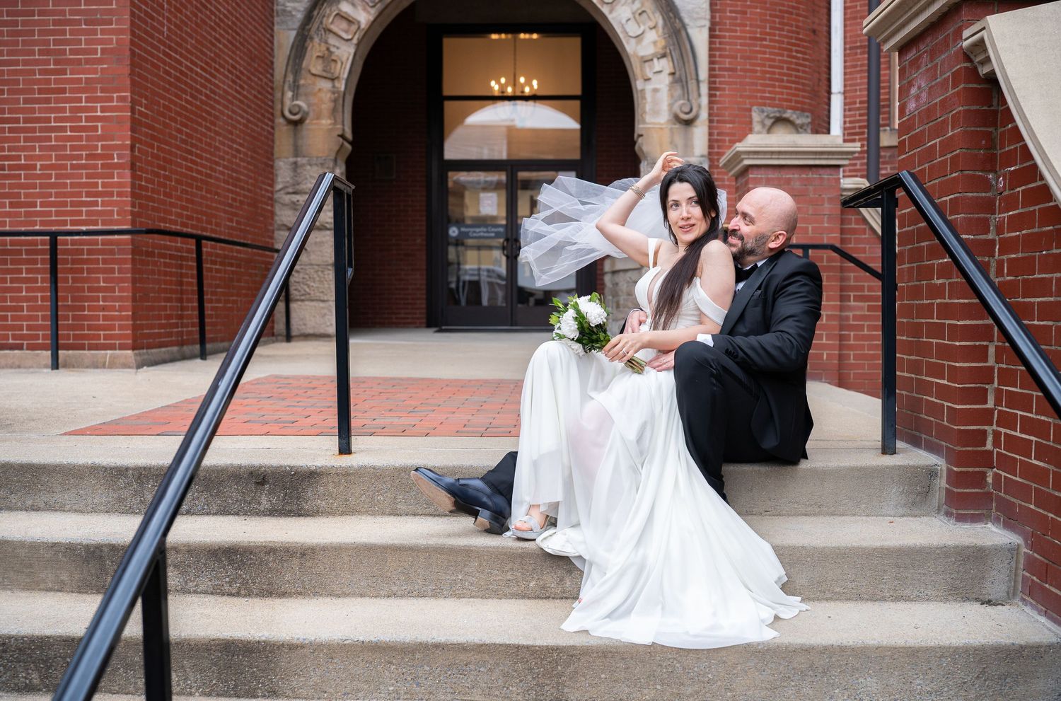 a bride and groom sit on the steps outside a red brick courthouse together, laughing as the wind blows her veil; shot in Morgantown, WV