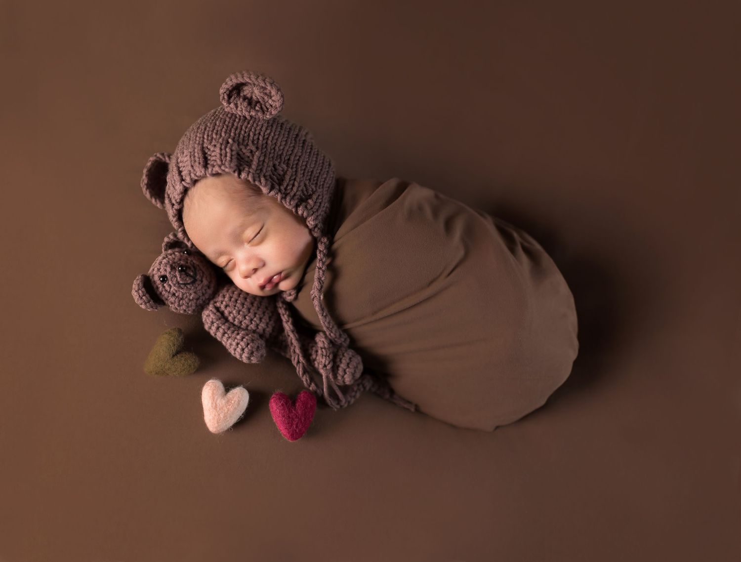 Newborn baby sleeping peacefully wrapped in brown swaddle wearing knit bear bonnet with small hearts on brown background.