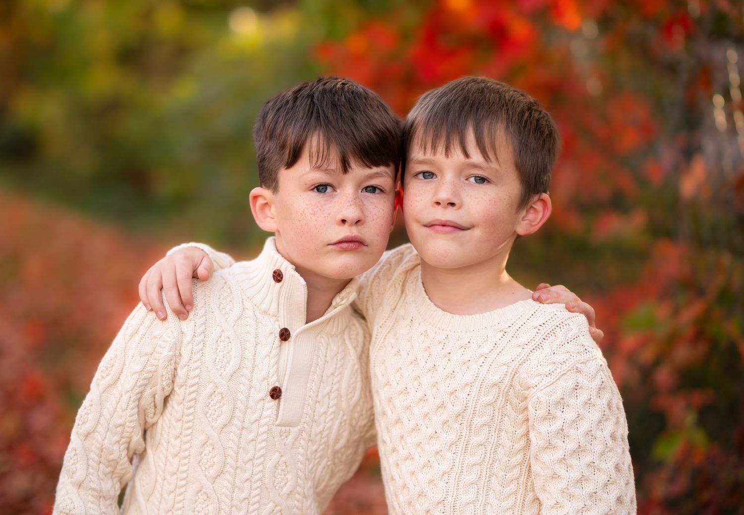 two young boys in ivory sweaters pose with arms around each other's shoulders for a portrait with bright fall leaves in the background