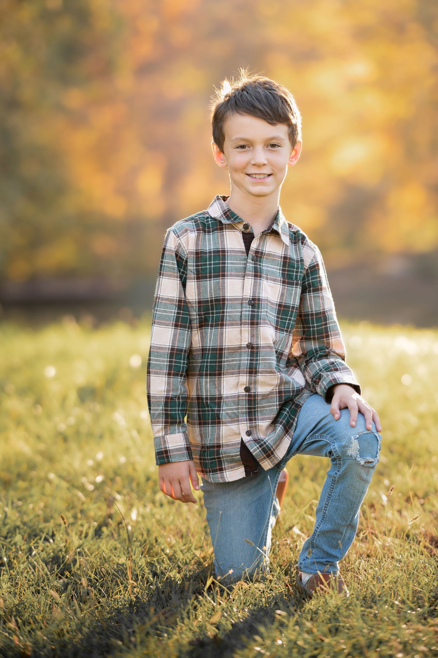 a young boy in a plaid shirt and blue jeans poses on one knee in the grass and smiles for a picture, with beautiful fall foliage behind