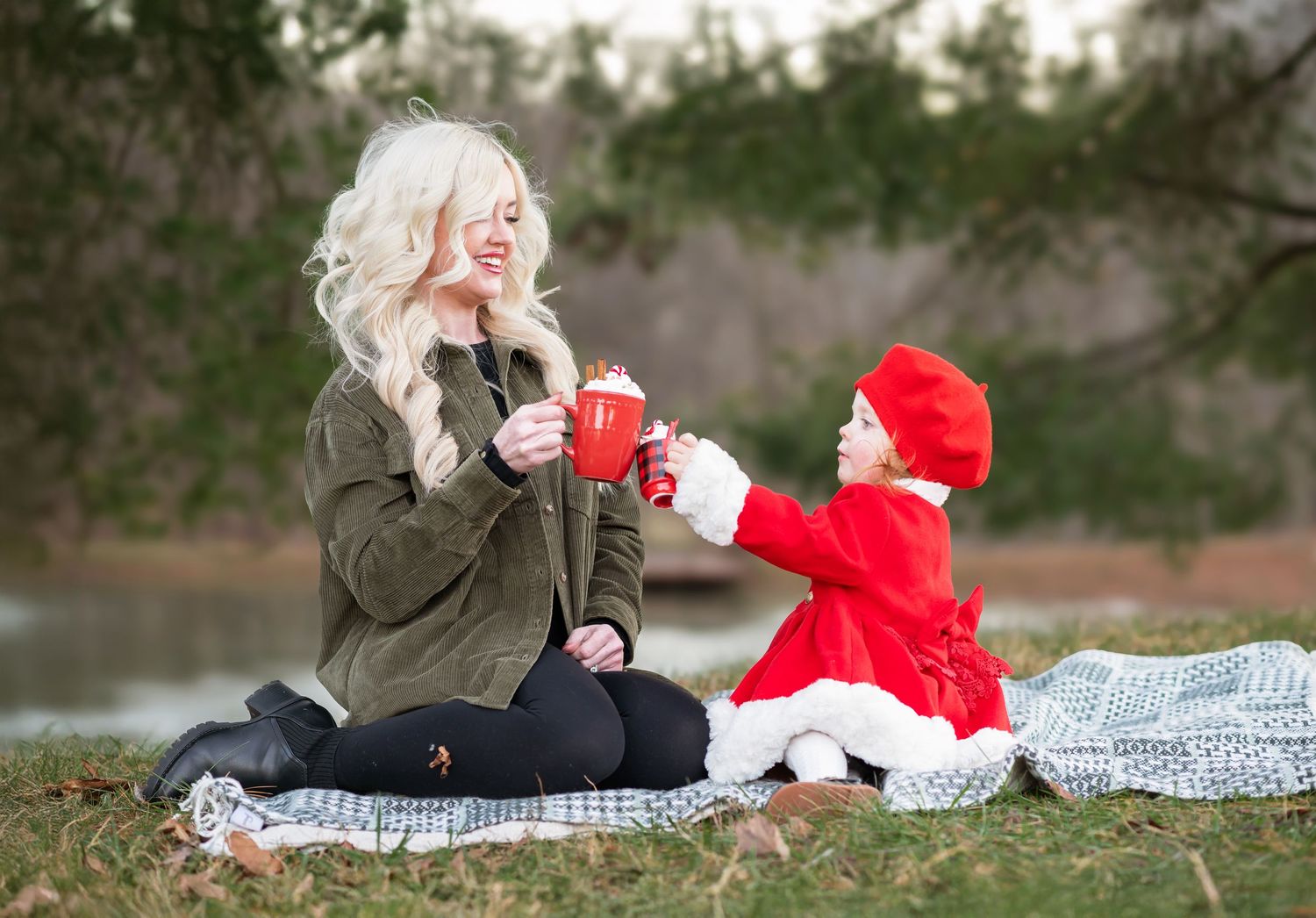 a blonde mother and daughter sit on a Christmas blanket in the grass with red mugs of hot cocoa and marshmellows