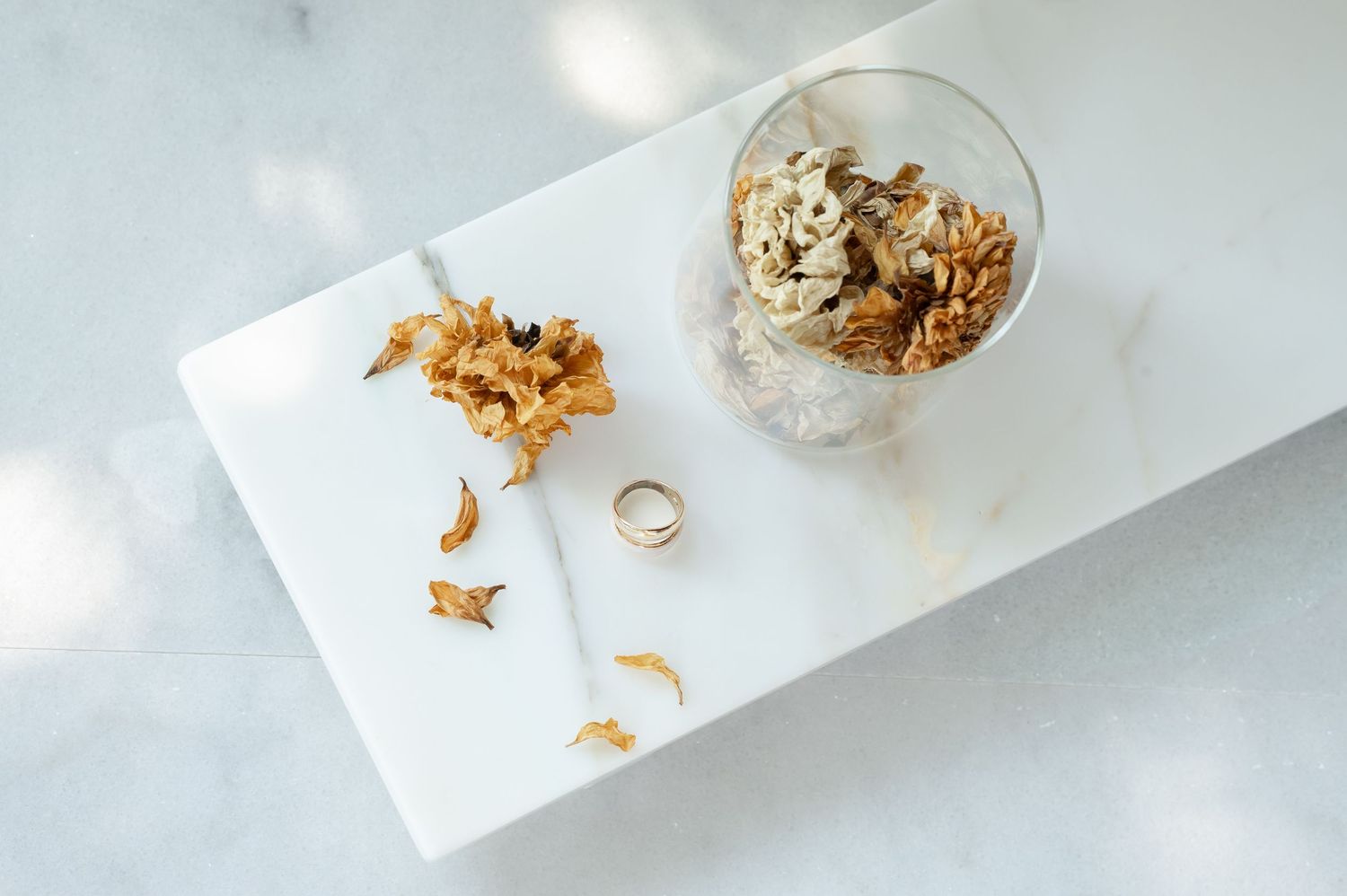 Gold wedding ring beside dried flowers and a glass jar of dried botanicals on a white marble tray