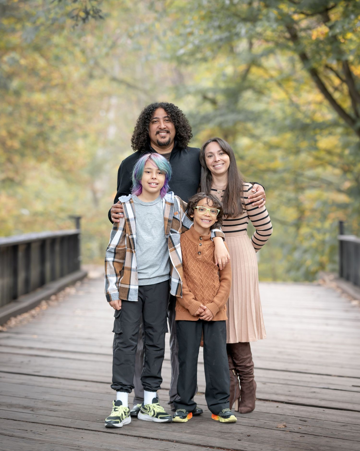 Family of four smiling together for a picture on a wooden bridge surrounded by autumn foliage in Valley Falls State Park, WV, taken by Laura