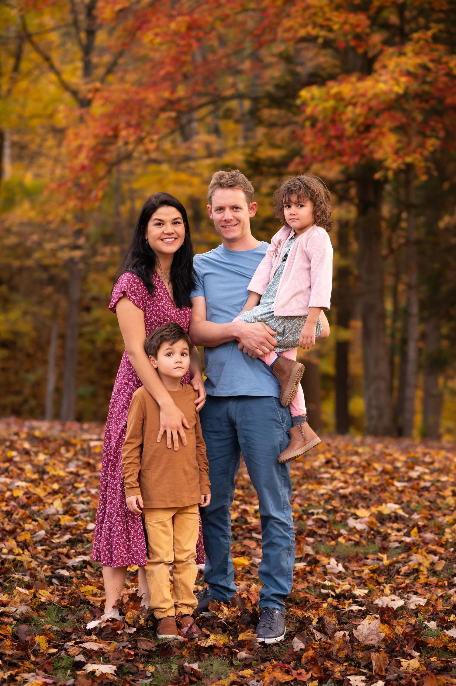 a young family with two small children stands in a park, surrounded by beautiful fall leaves, and poses for a portrait