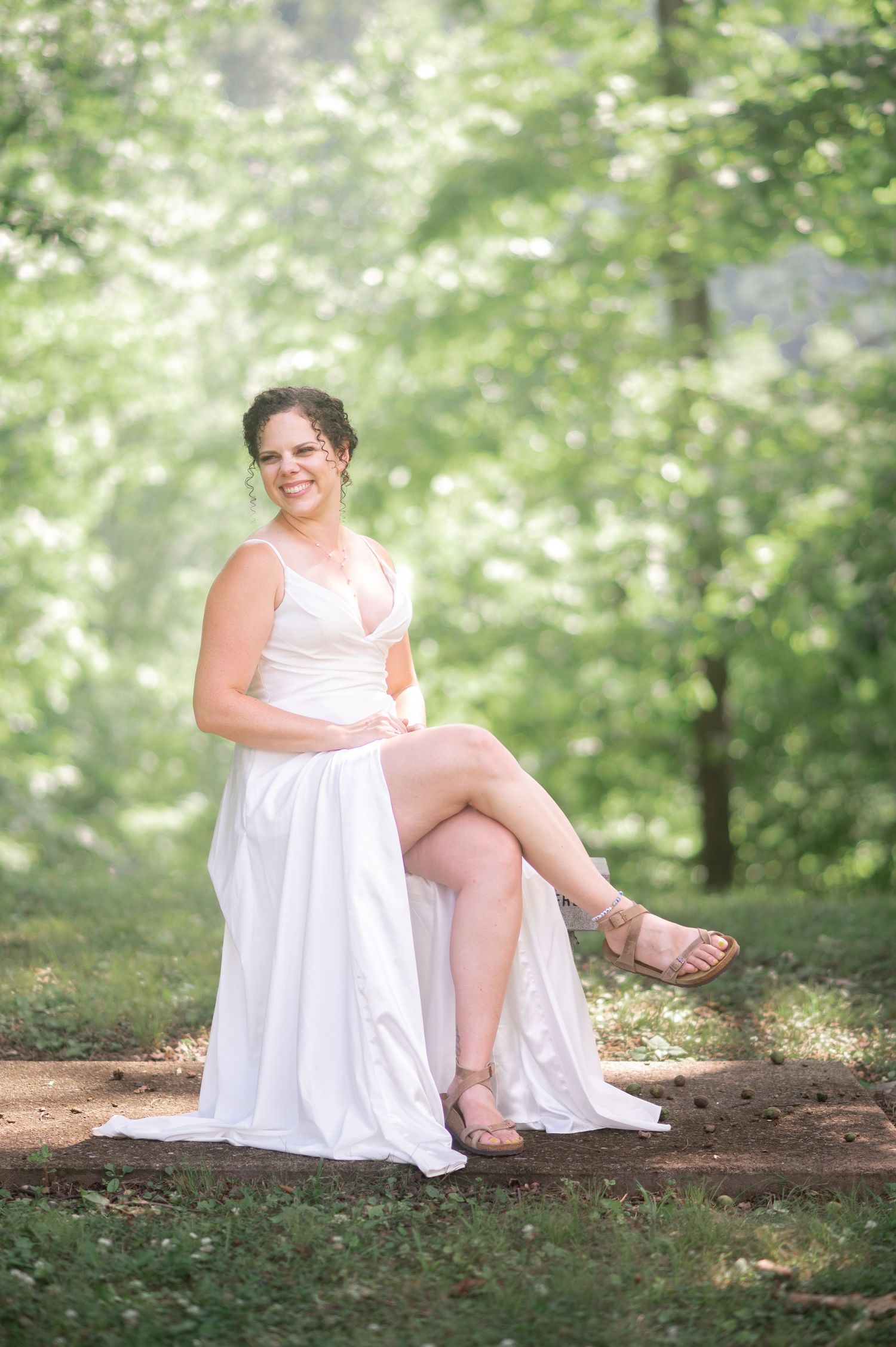 natural outdoor portrait of a dark-haired bride, seated and smiling with sunlit trees in the background