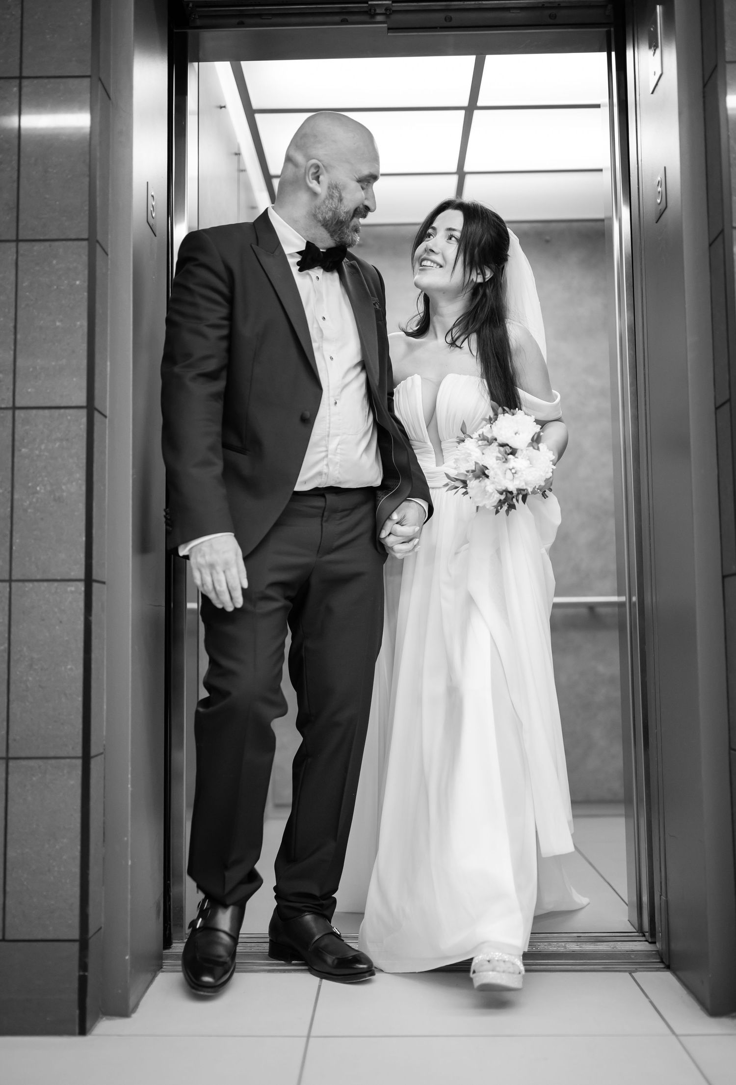 a bride and groom step out of an elevator together after their wedding ceremony, laughing