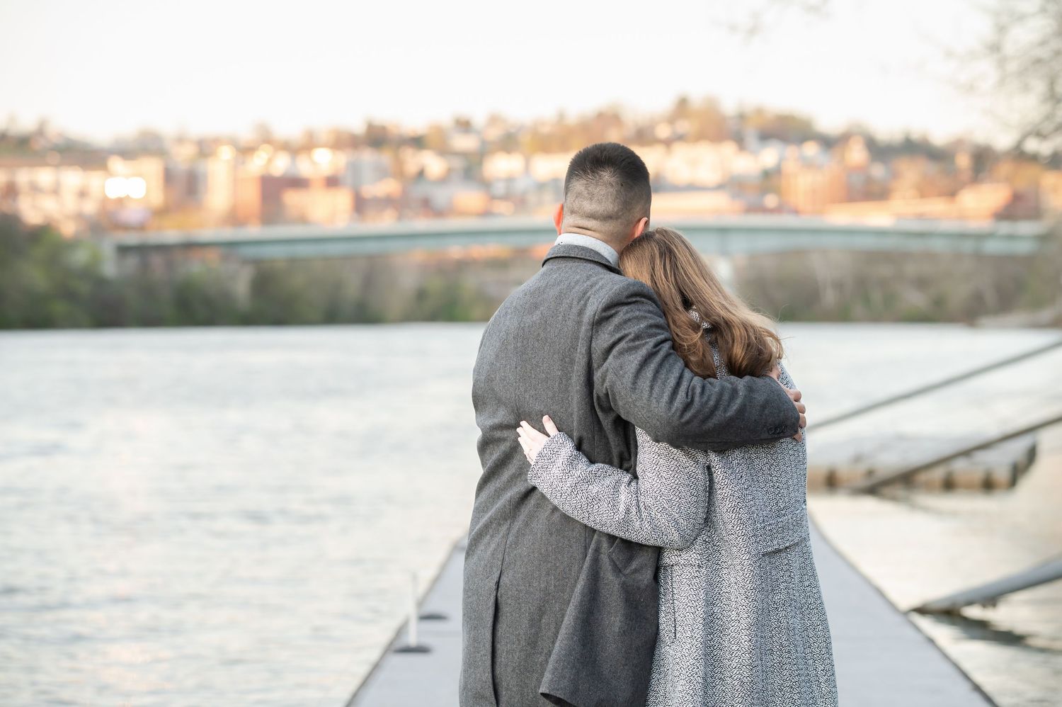 a couple in gray coats embraces as they stand on a dock by the river looking over the city