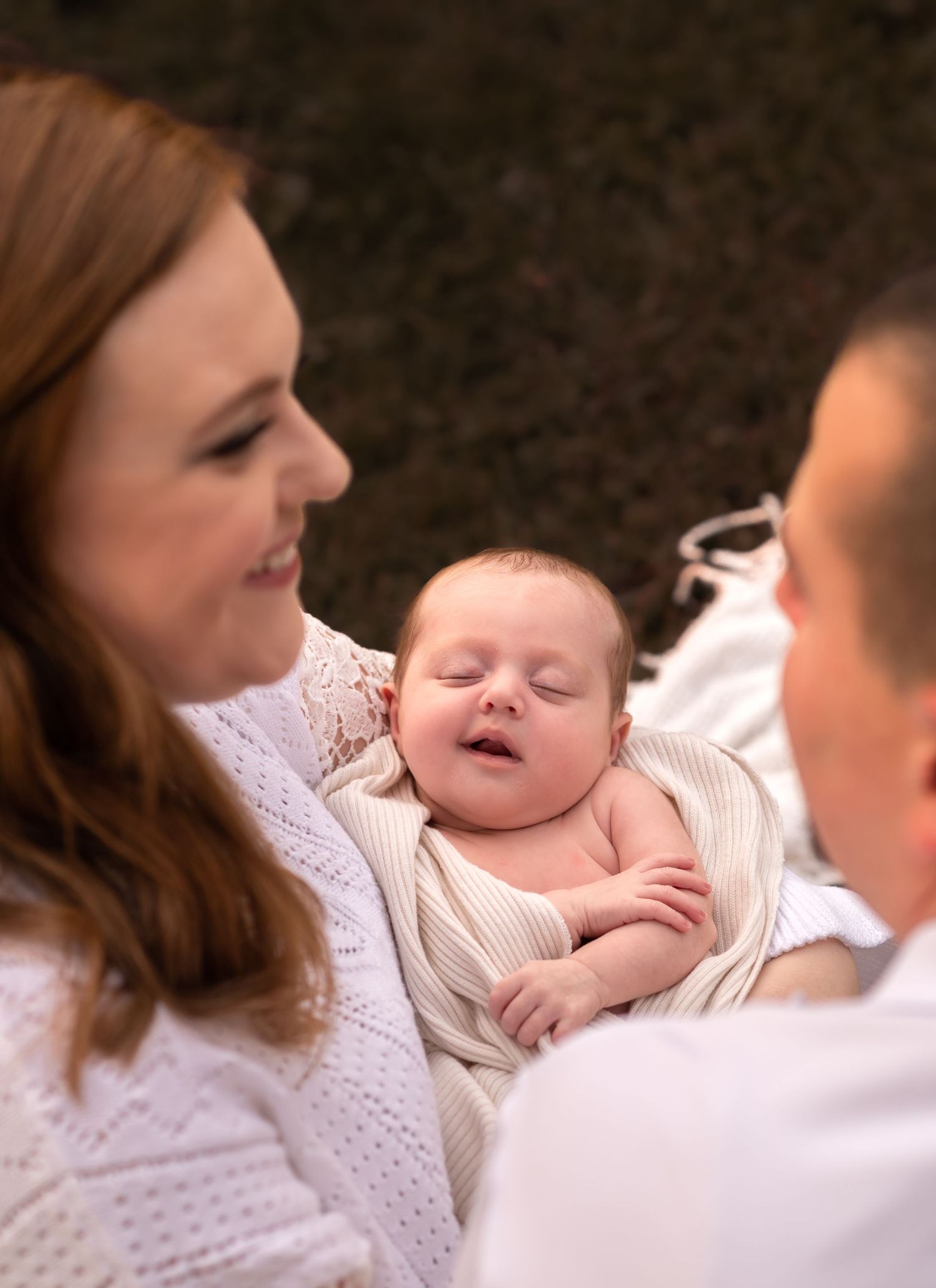 Parents gazing lovingly at their sleeping newborn baby wrapped in a white swaddle during an outdoor family photo session.
