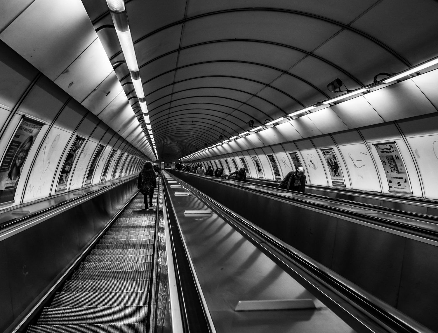 Black and white photo of a long underground subway escalator tunnel with fluorescent lighting and a lone figure.