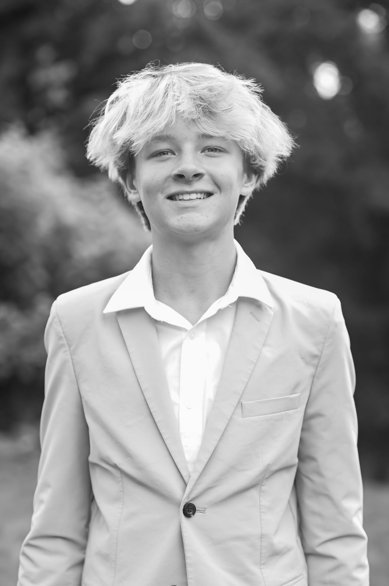 black and white portrait of a young man in a suit jacket, smiling for the camera with trees in the background