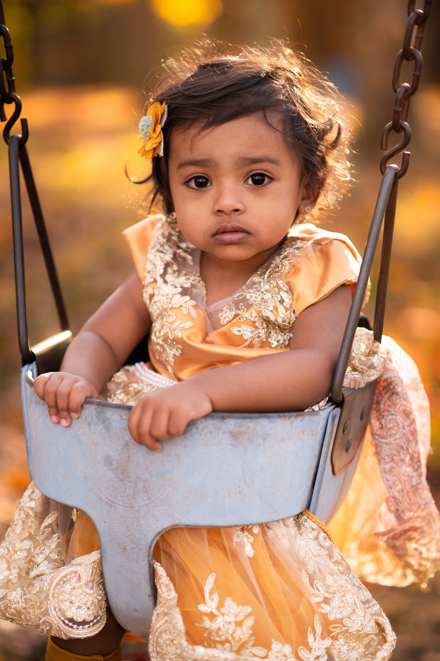 a toddler girl with dark hair and dark eyes, in a lacy bright yellow dress, sits in a swing