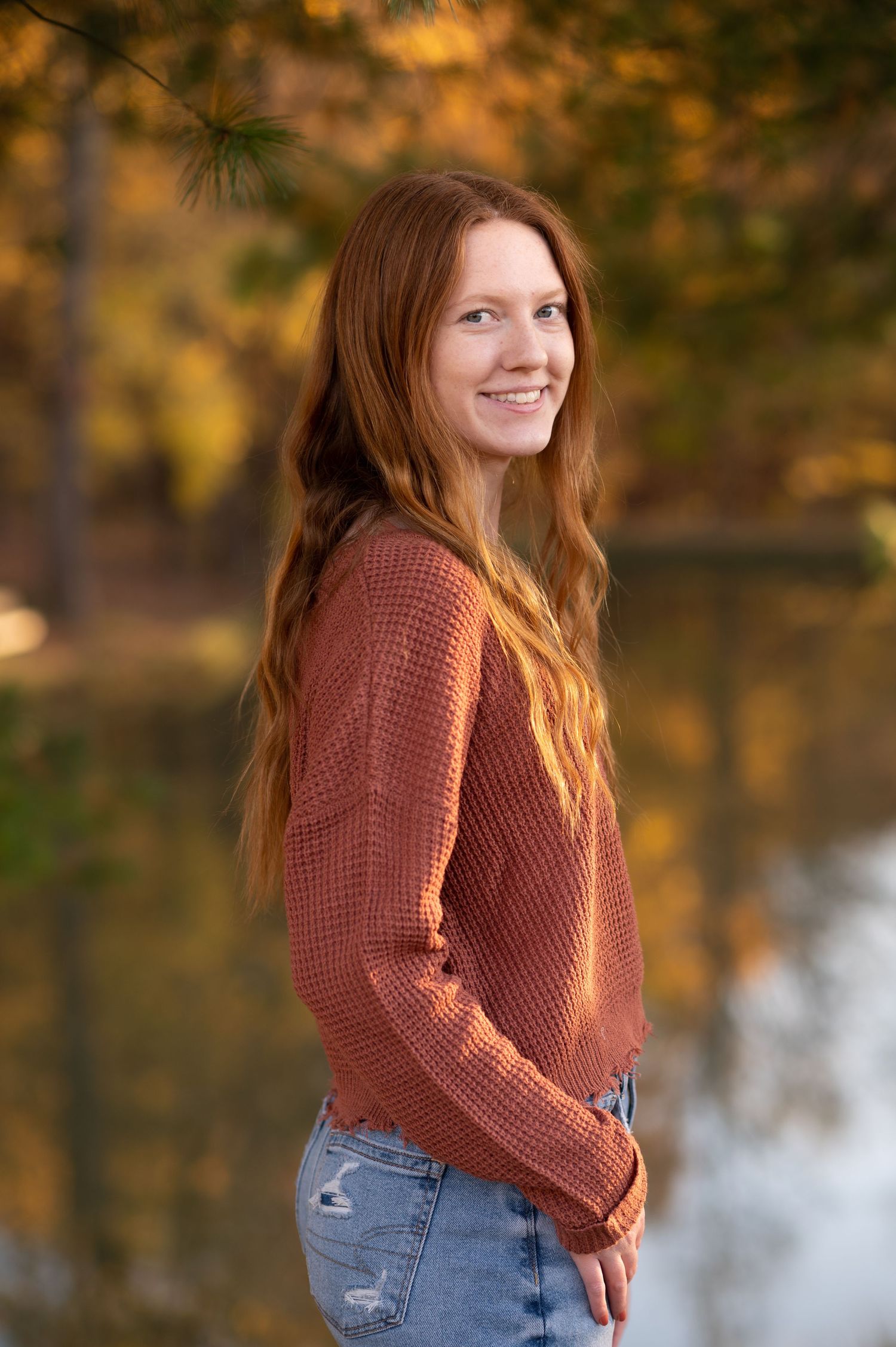 a pretty red-headed young woman in jeans and a rust colored sweater poses with a hand in her pocket for a senior picture
