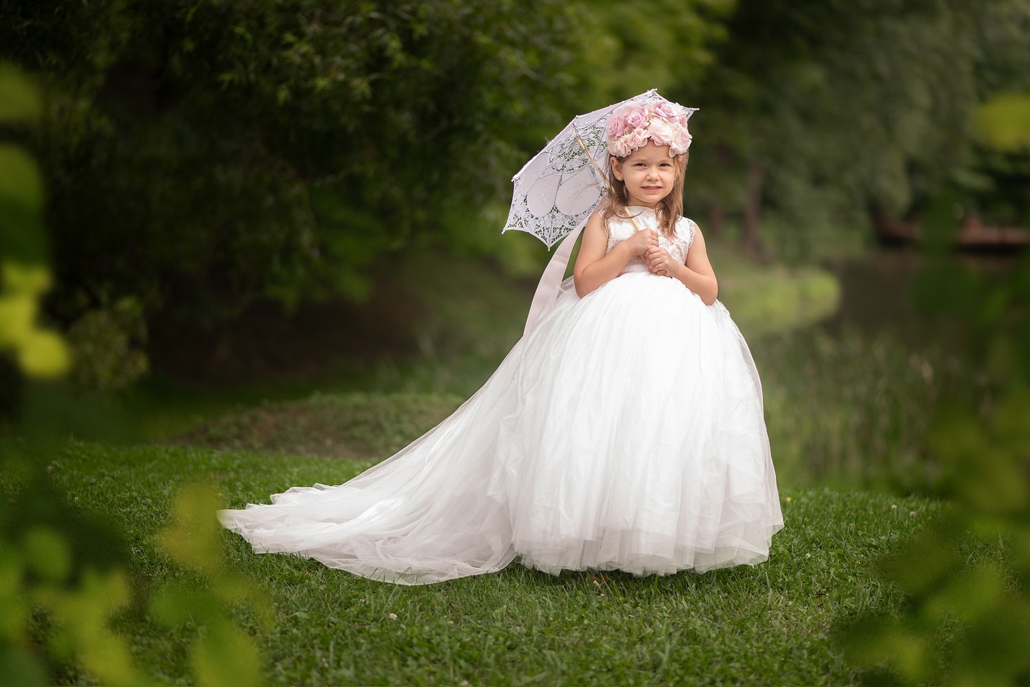 a little girl in a floor length white gown stands with a white parasol in the fluffy green grass with trees around