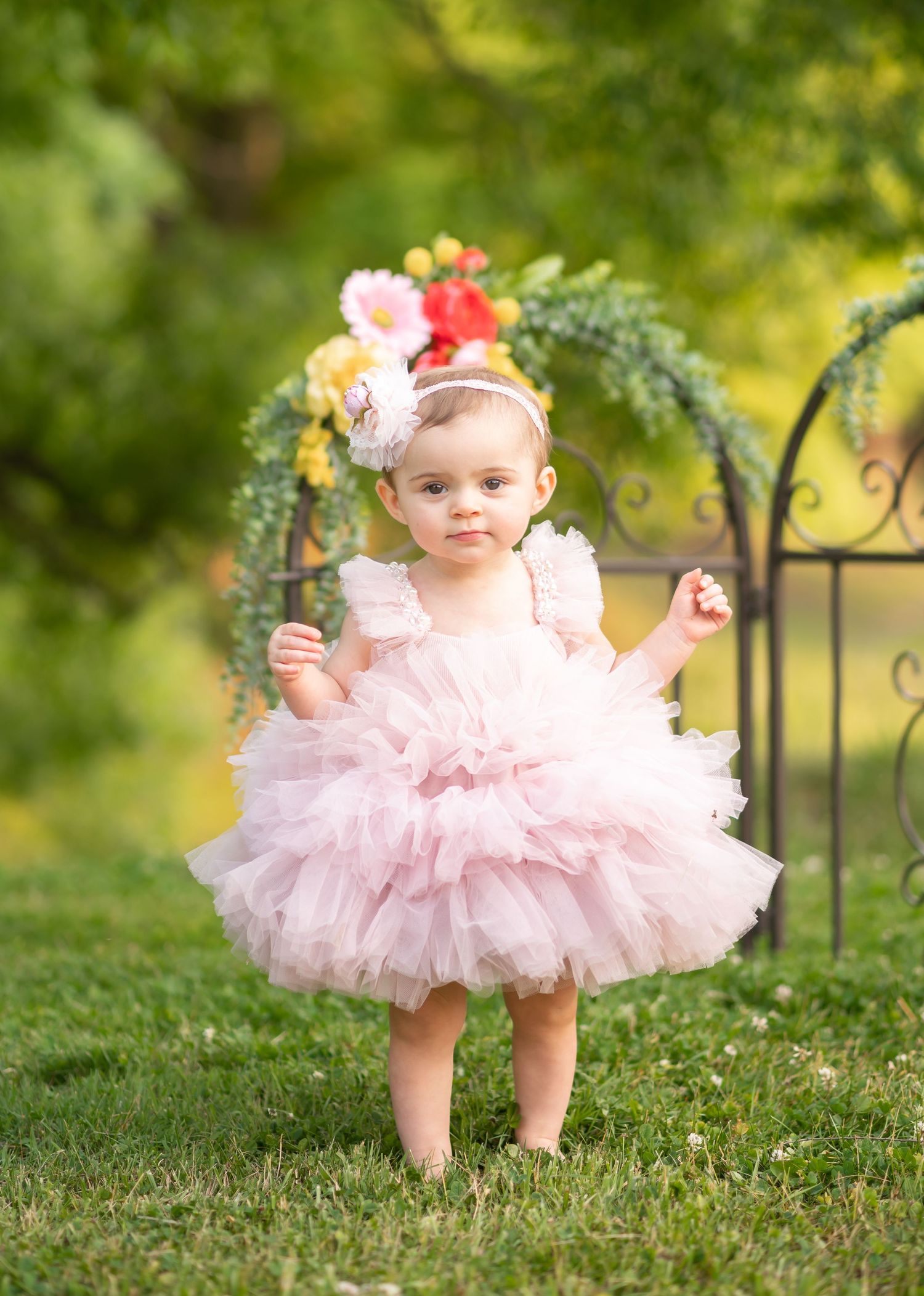 Birthday portrait of a one year old girl in a fluffy frilly pink dress, standing in the grass with a floral arch and trees in the background