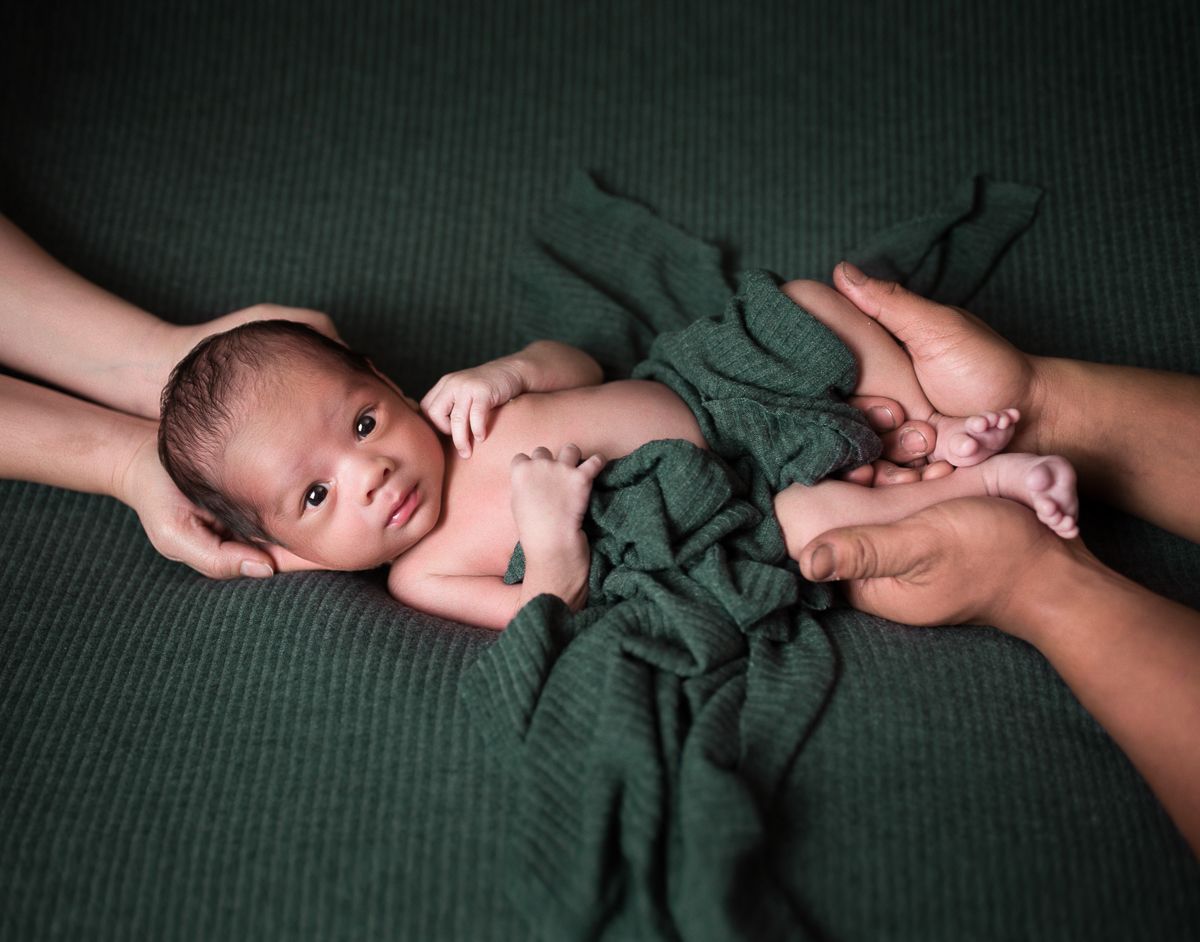portrait of a newborn baby boy being held by his father's and mother's hands, against a rich dark green background