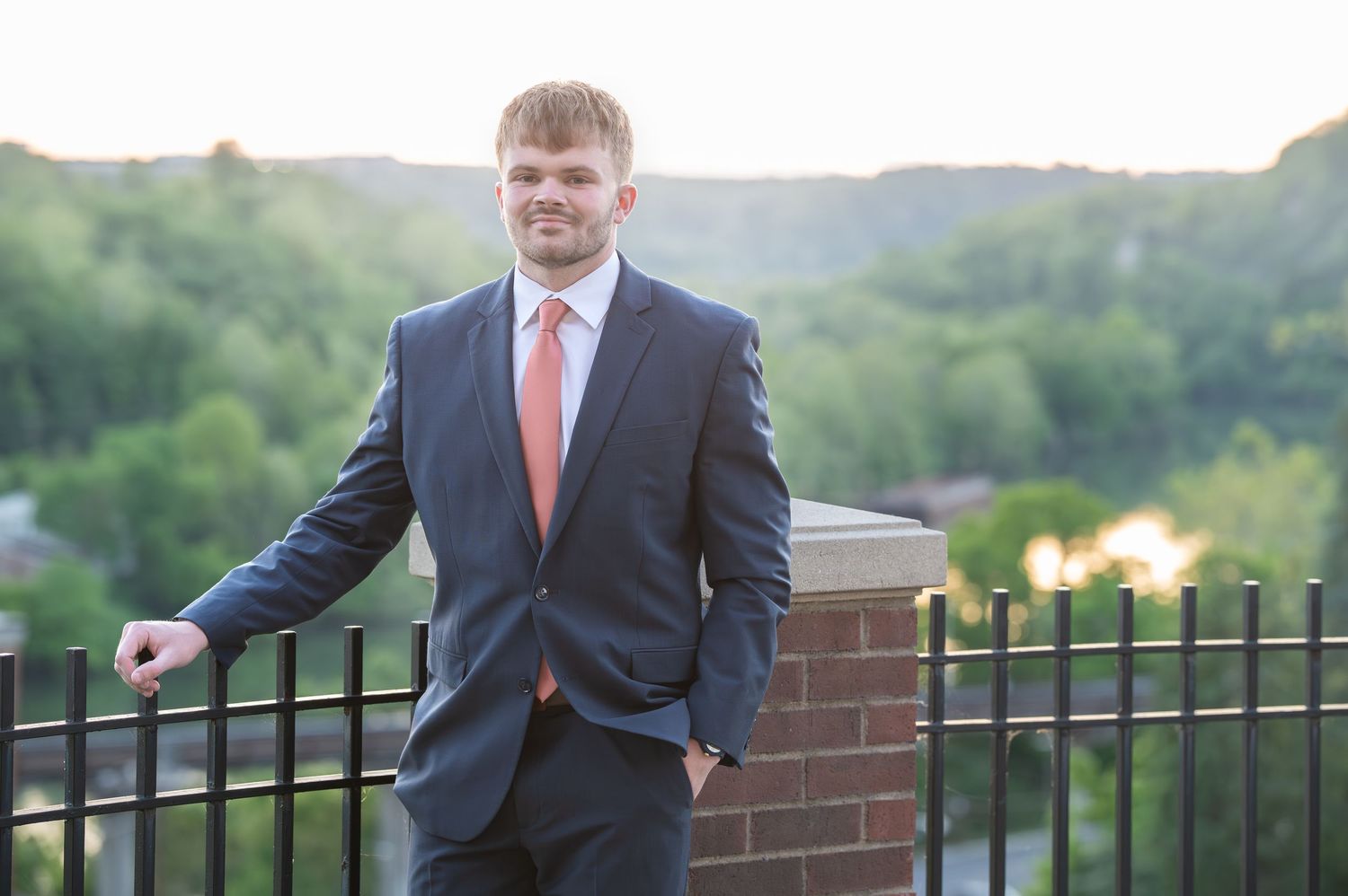 A WVU graduate in a blue suit and red tie leans against a railing with the Monongalia River in the background in Morgantown, WV
