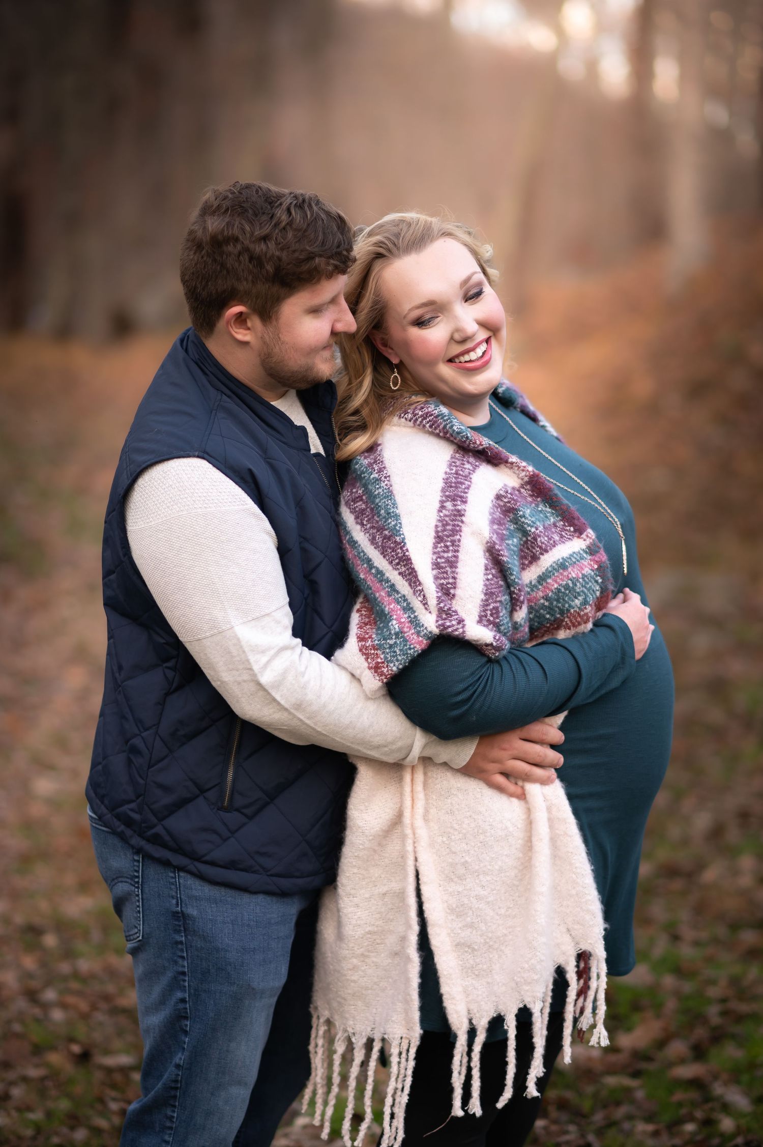 a young blonde pregnant woman and her husband embrace and laugh for a portrait with rich autumn foliage behind them in Morgantown, WV