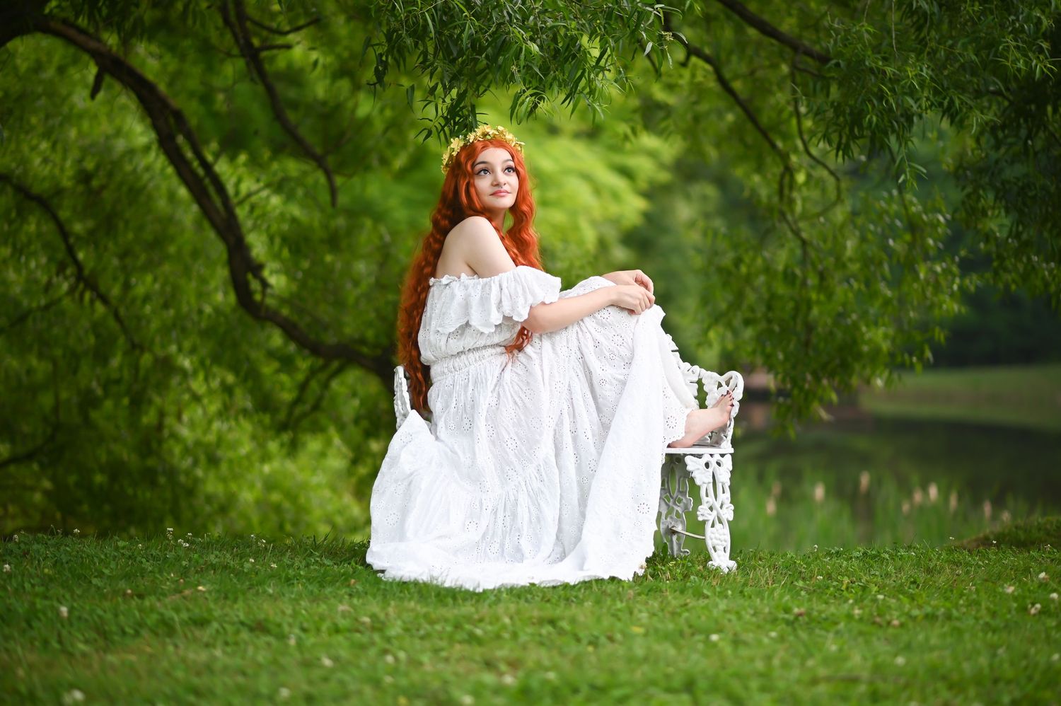 a beautiful young woman in a flowing white dress with very long red hair poses on a white antique bench with lush green trees and grass