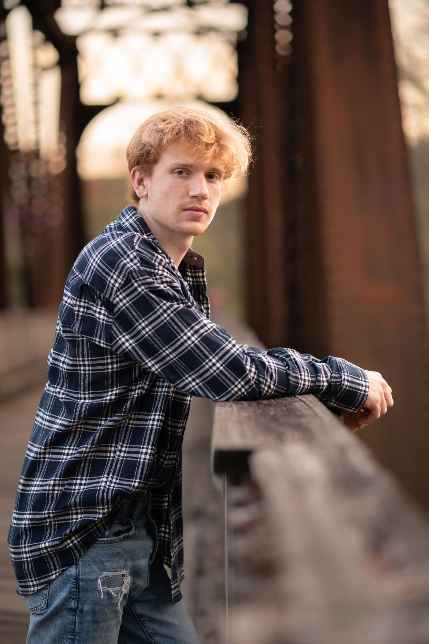 a curly haired boy in a plaid jacket and solemnt expression leans against the railing of an old wood and iron bridge in downtown Morgantown