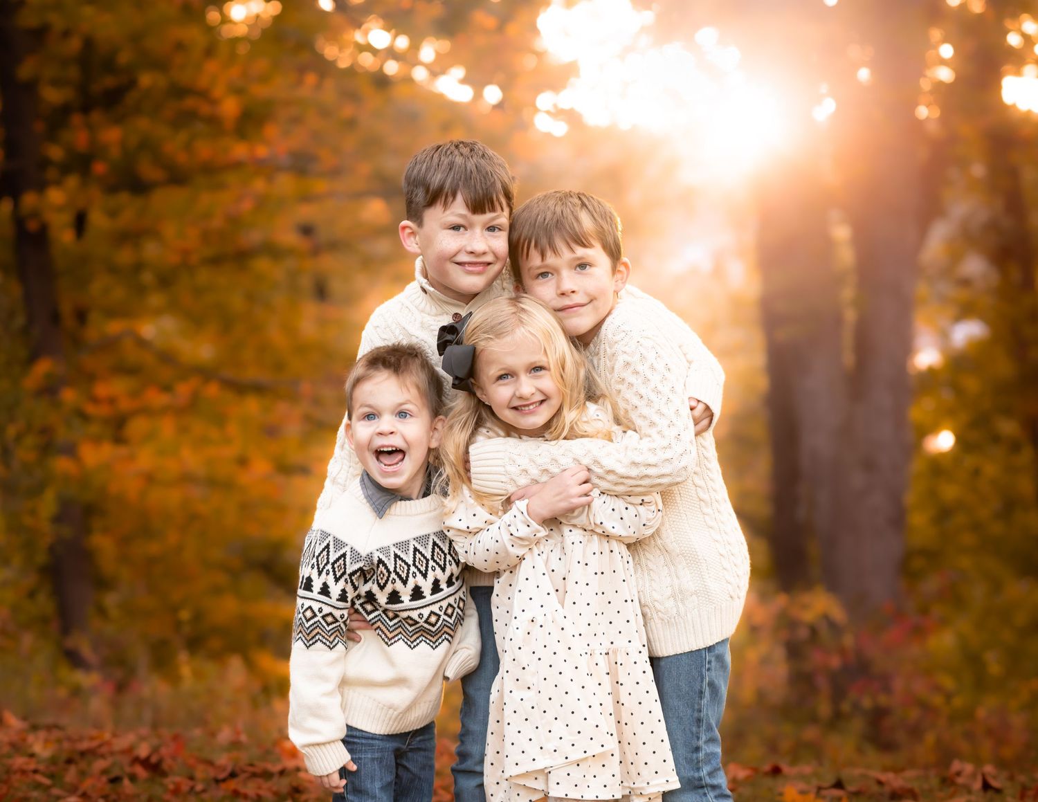four siblings in matching ivory sweaters huddle close together and smile for a picture, with brilliant fall foliage in the background