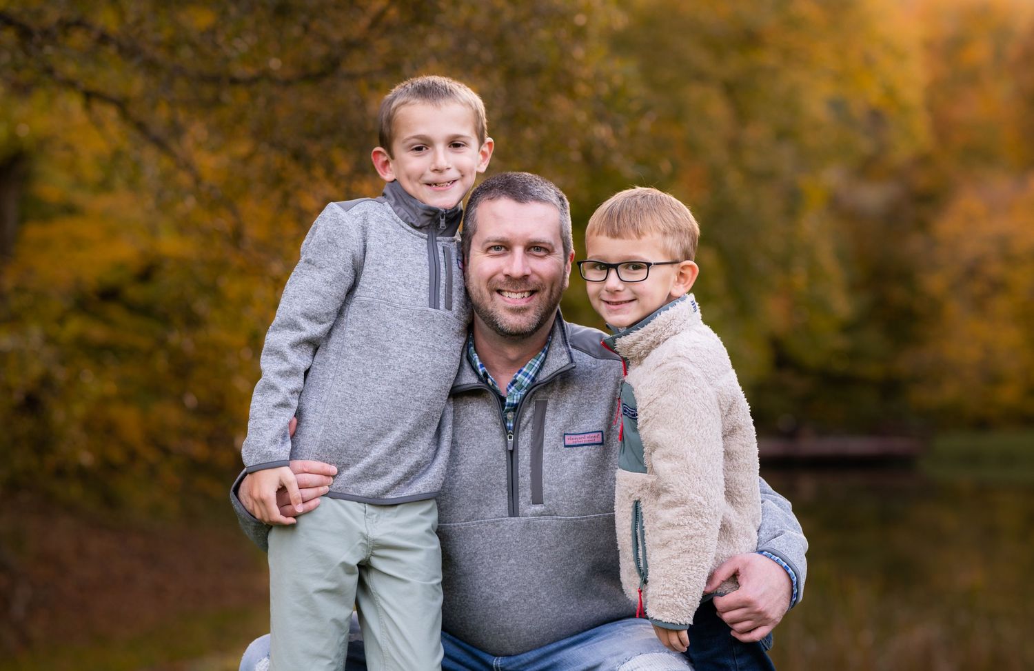 a dad crouches down to put his arms around his two young sons for a portrait with beautiful fall leaves in the background