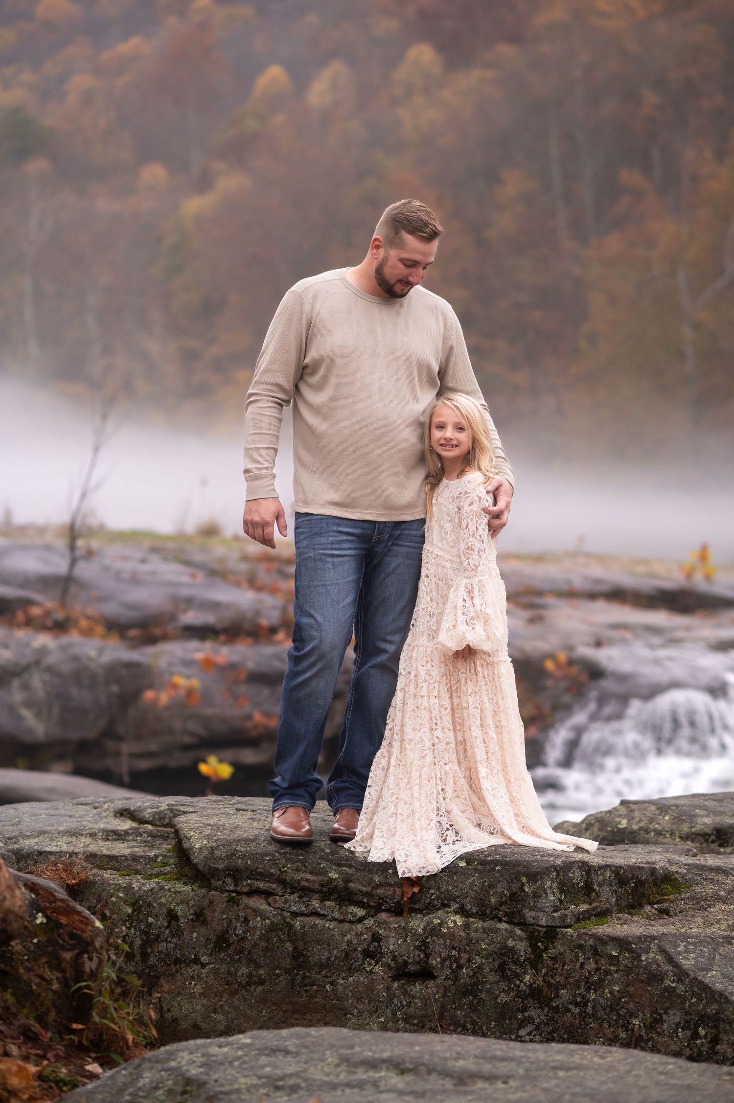 a father and daughter pose together on a riverbank with fog and fall leaves in the background