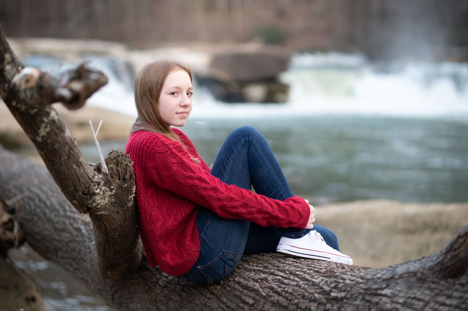 a young student in a red sweater and jeans sits curled up on a large fallen tree with a roaring river and waterfall in the background