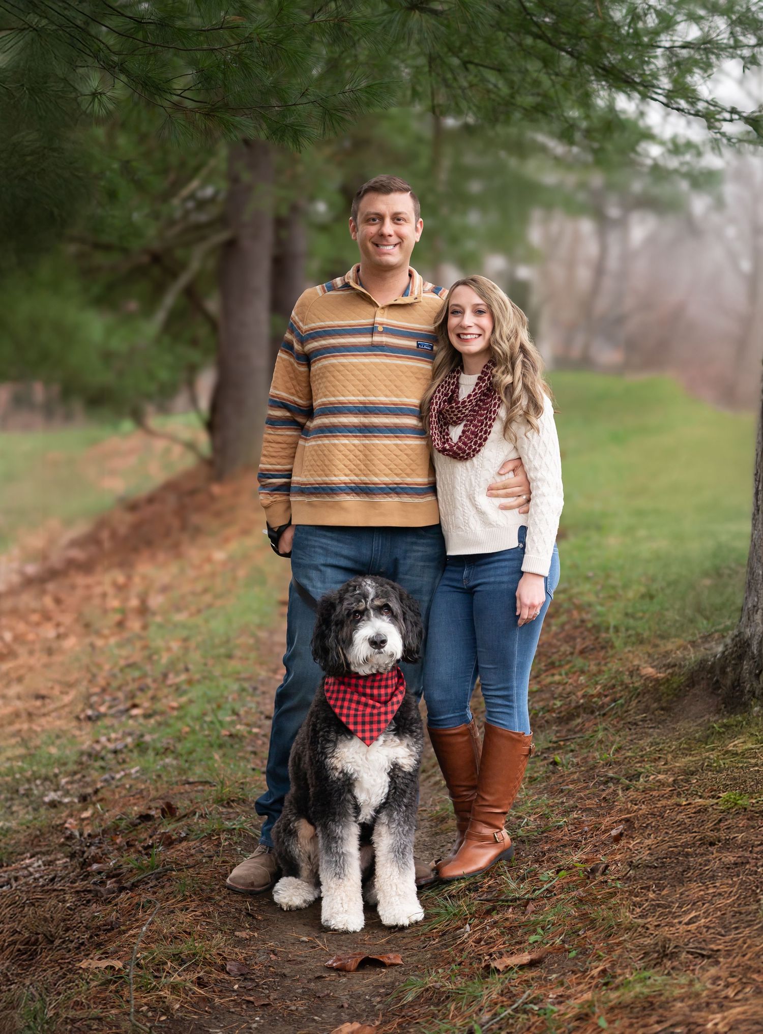 a sweet young couple and their dog pose together and smile for a Christmas card picture in front of evergreen trees