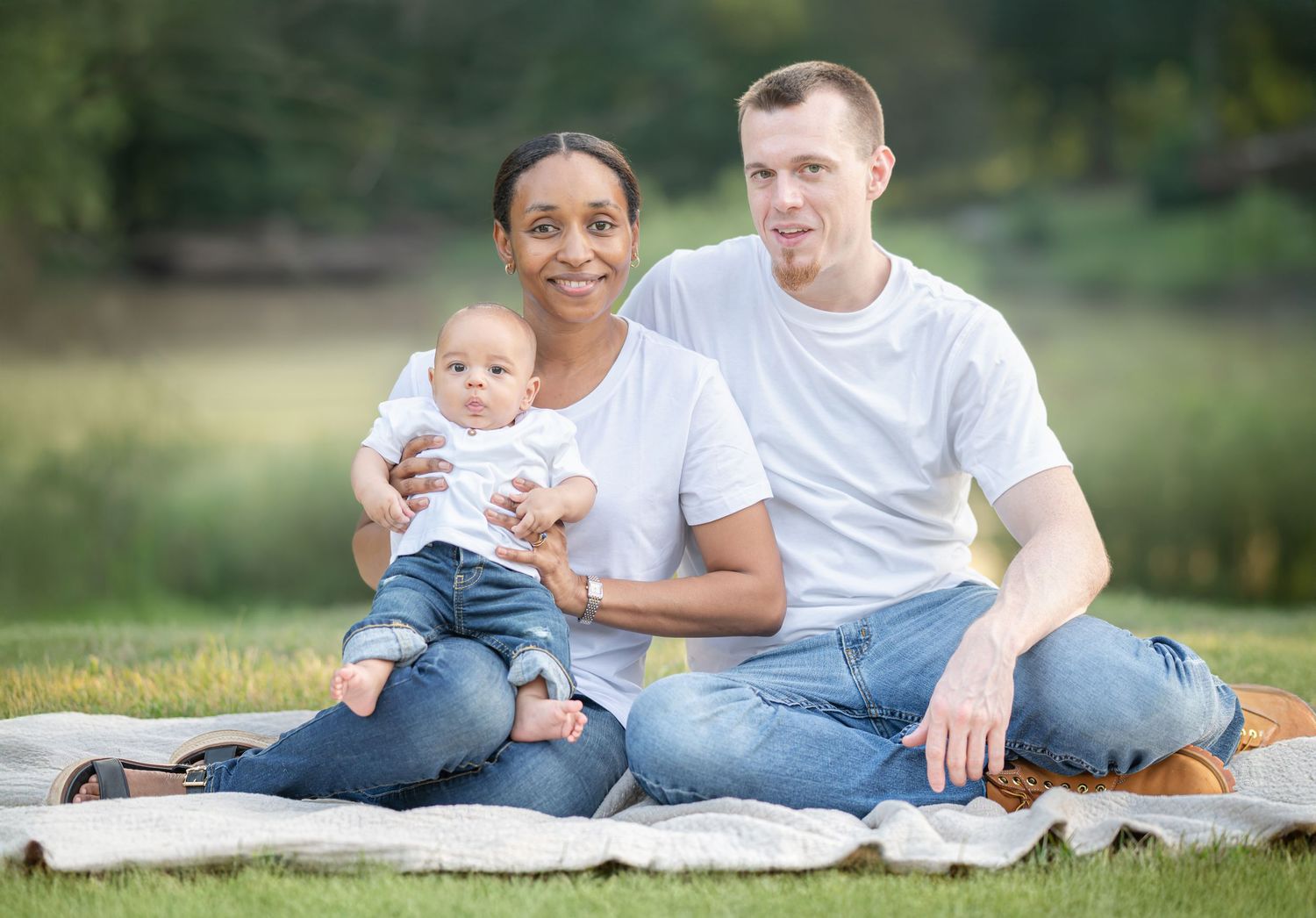 a young husband and wife pose with their baby boy on a blanket on the grass, with a pond and trees in the background, in Morgantown, WV