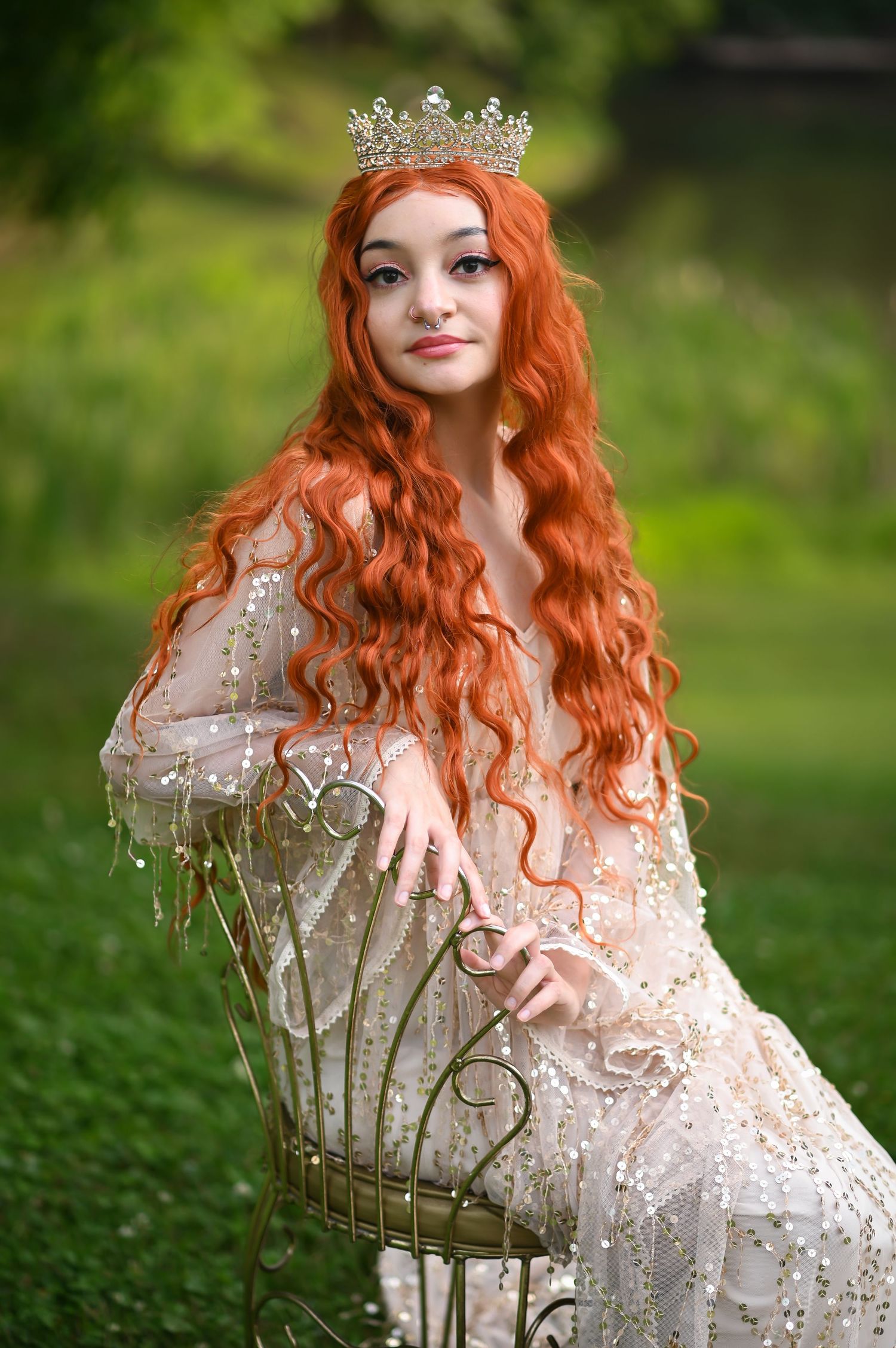 a young woman with long curly red hair in a gold sequin dress and crown sits in a gold chair in the middle of lush greenery outside