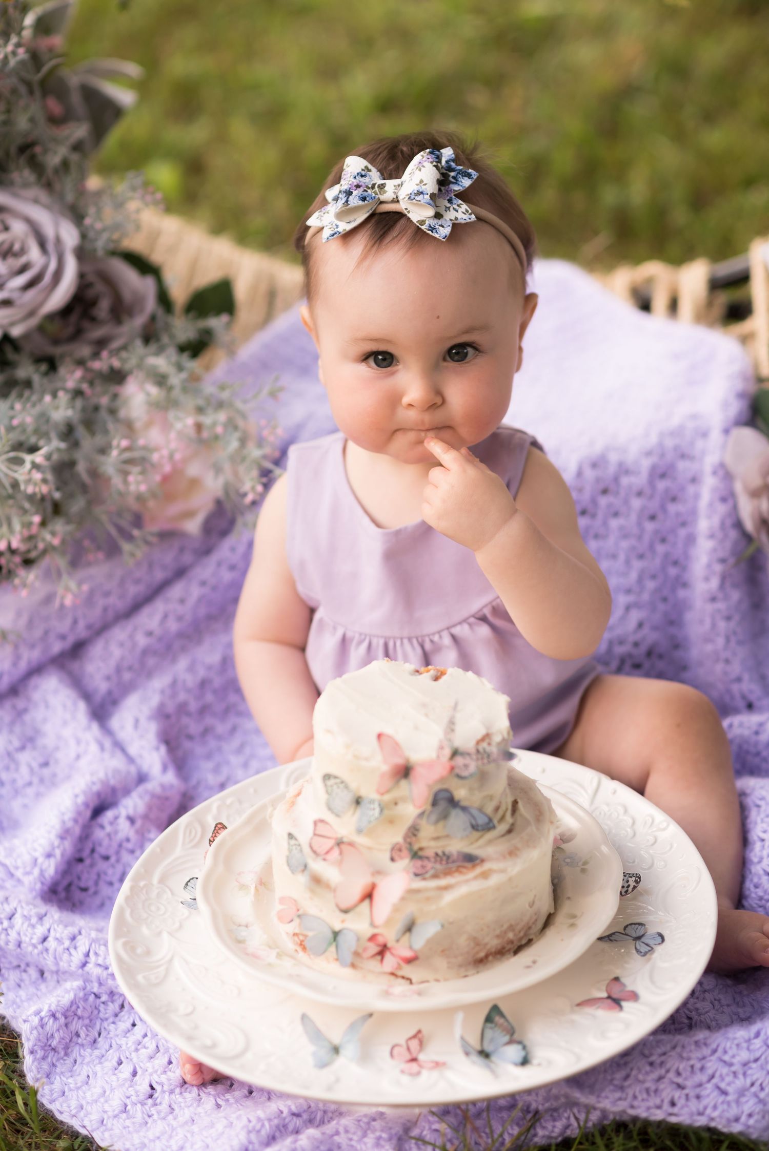 a little girl dressed in purple sits on a purple blanket with a butterfly themed cake in front of her, under an arch of spring pastel flower