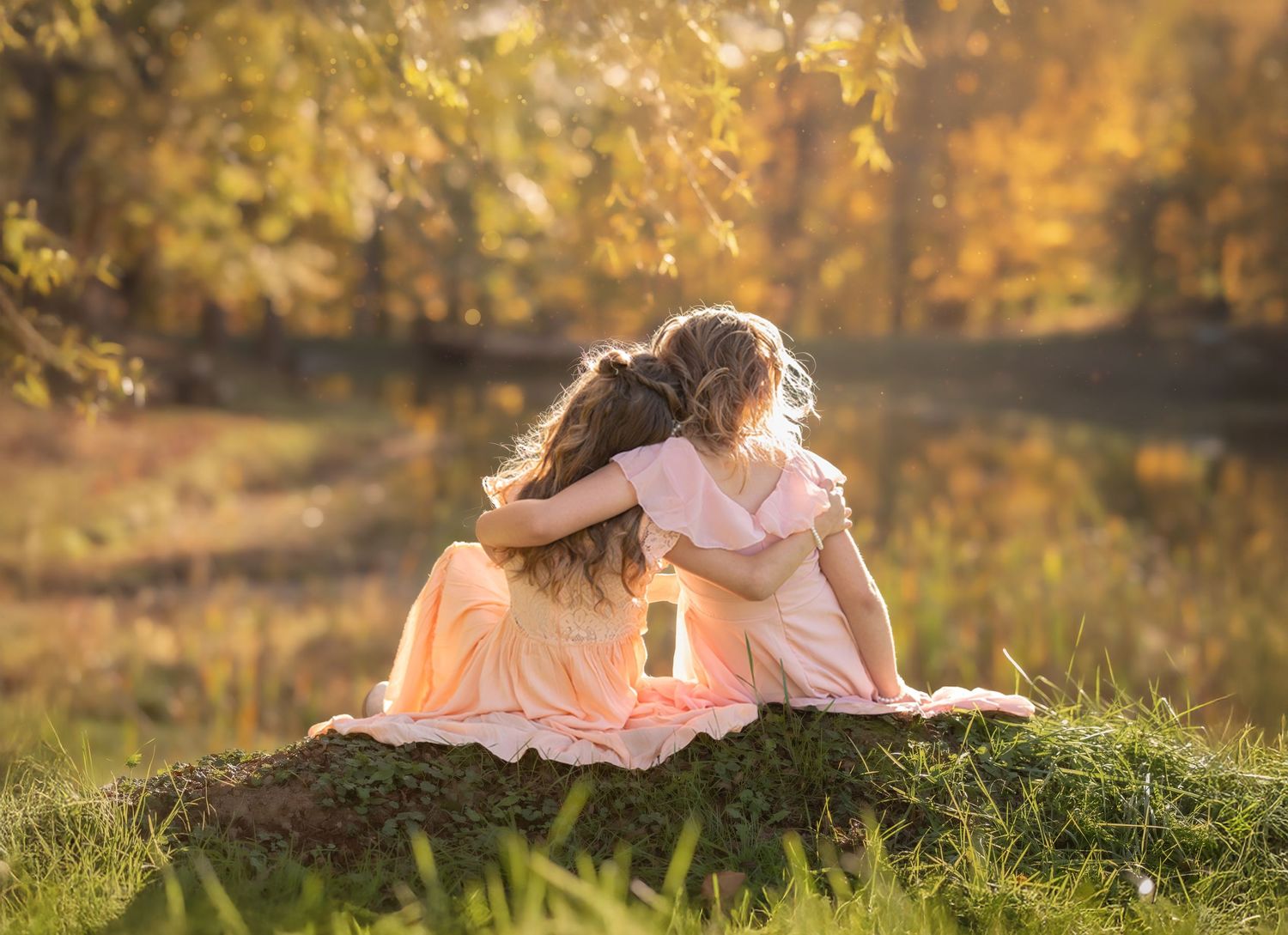 two little girls in pink dresses sit with arms around each other, looking over a beautiful sunlit pond and fall trees with golden glow