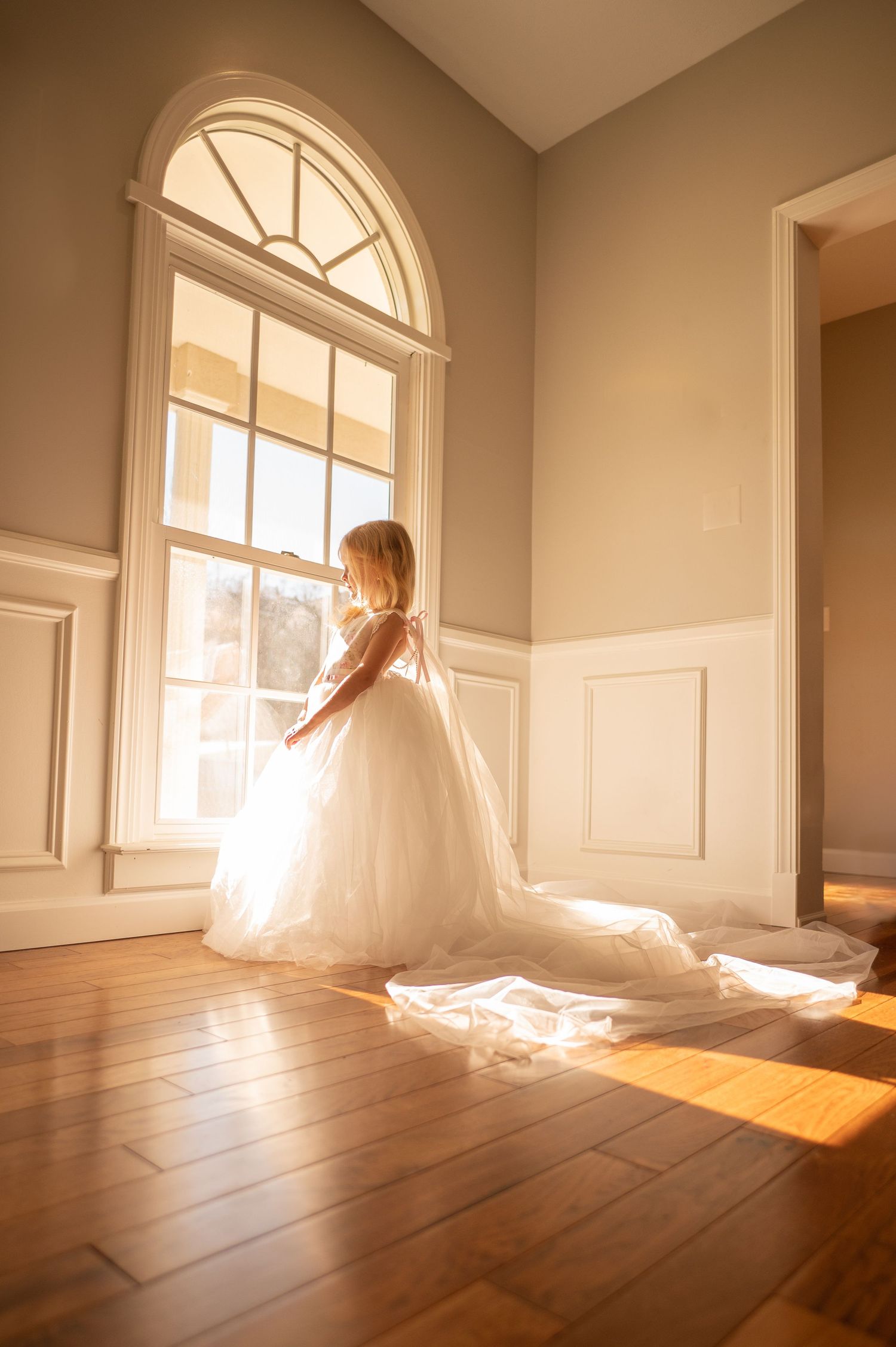 a little girl in a floor length white gown stands in front of a tall arched window in a sunlit room, looking out over the outside scenery