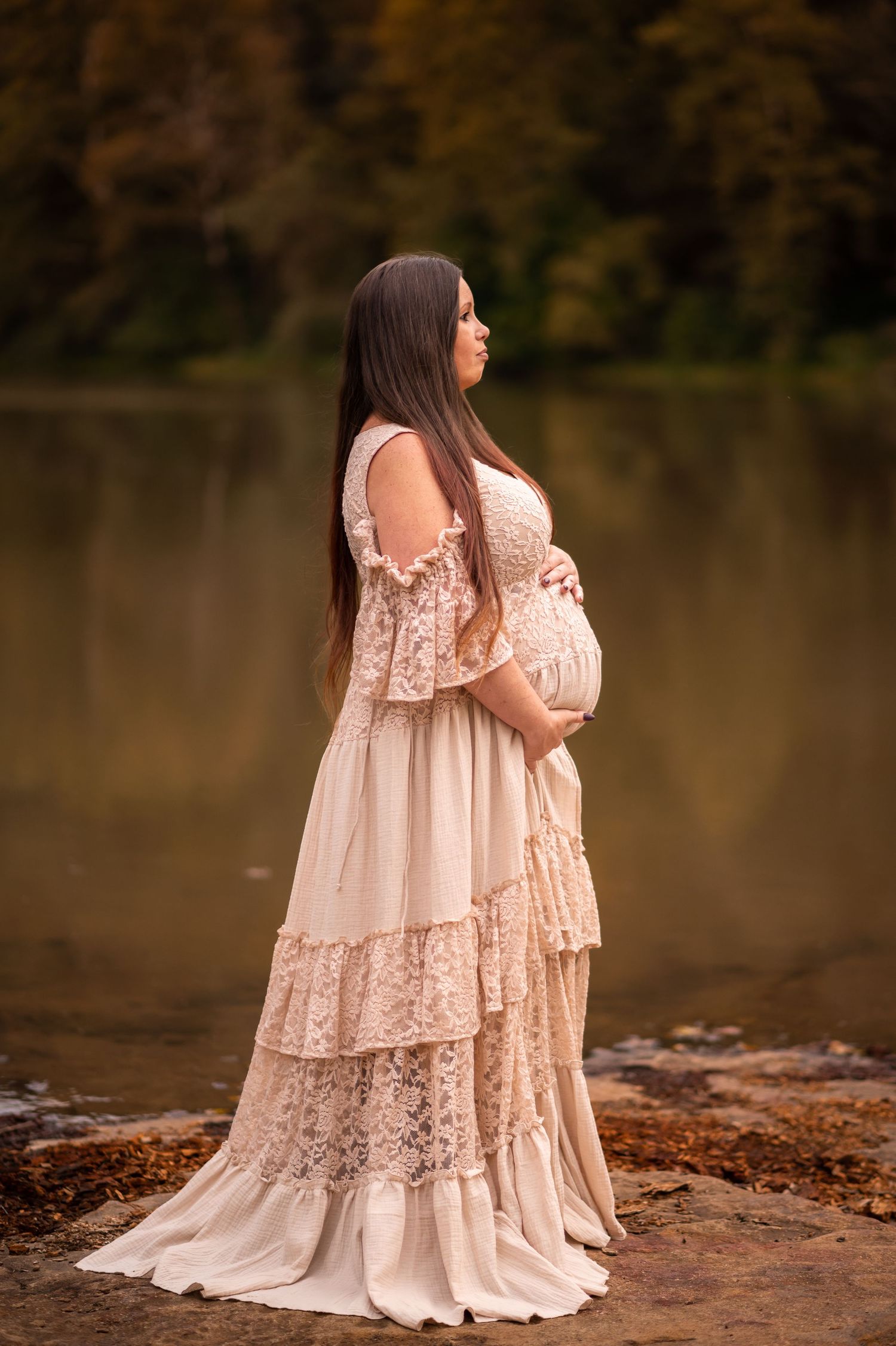profile portrait of a lovely pregnant woman with dark hair in a long beige lace dress, showcasing her pregnant belly with a lake behind her
