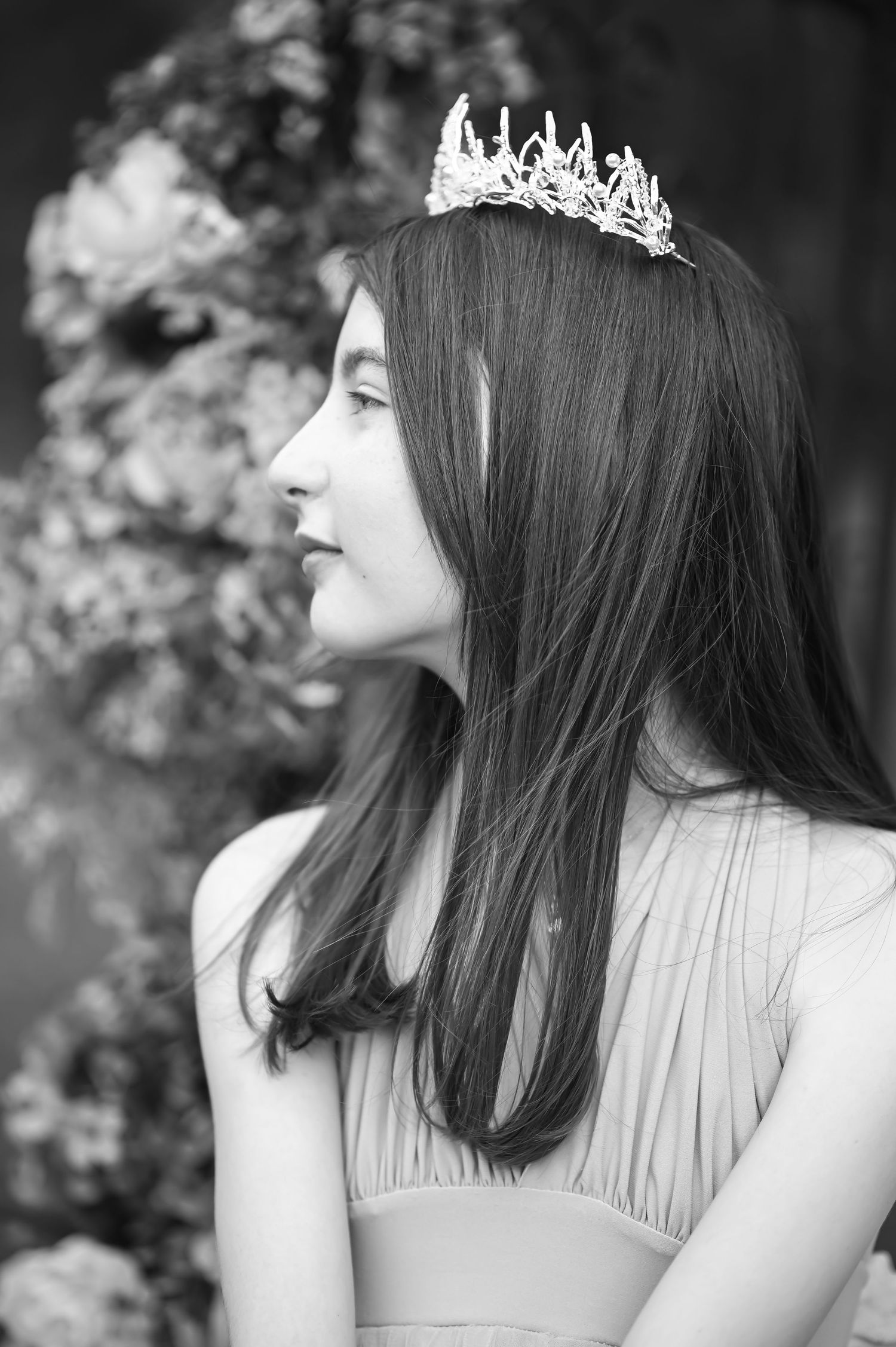 black and white closeup portrait of the profile of a dark haired young woman in a dress and tiara, with flowers in the background