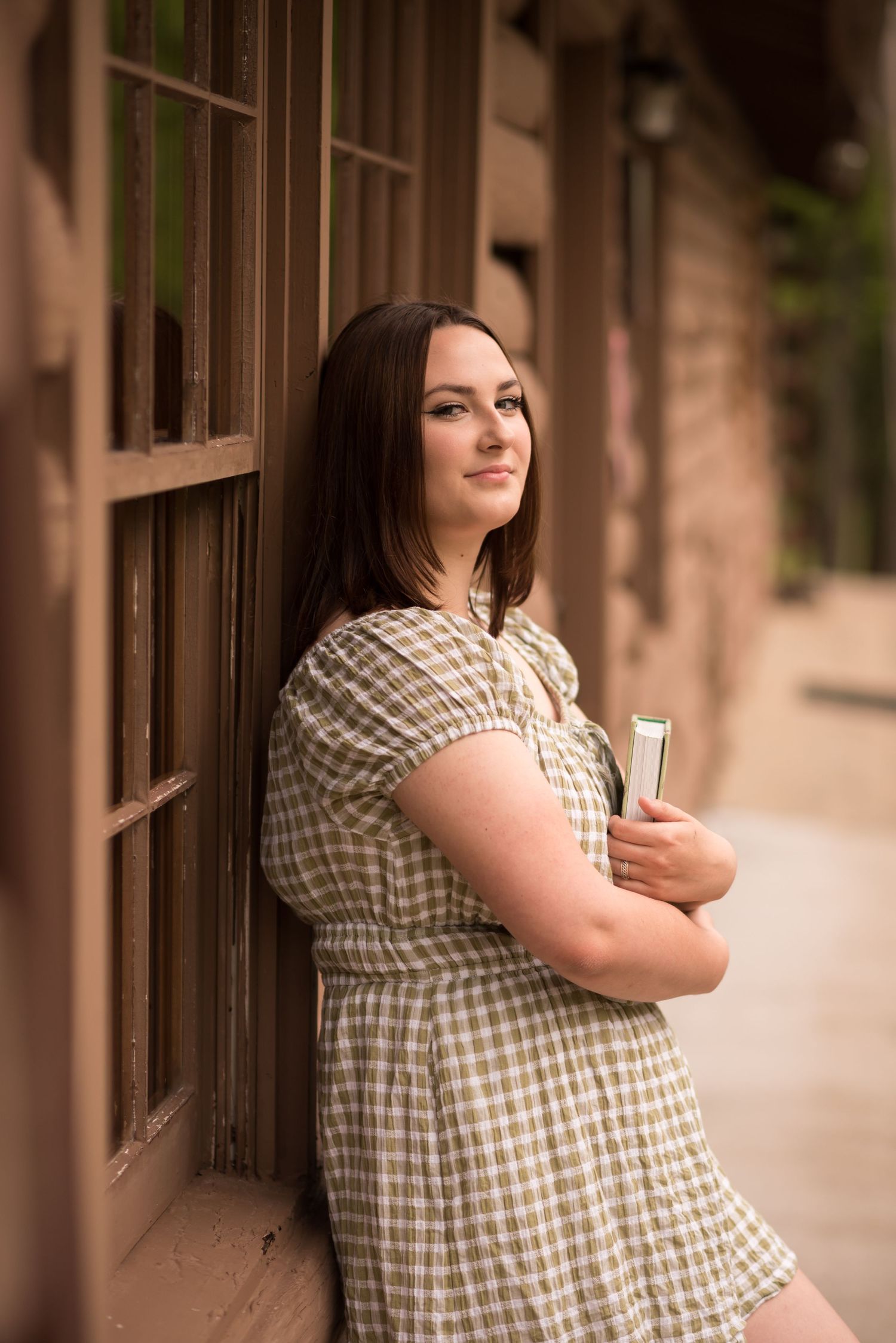 a dark haired high school student in a green dress leans against an old wooden window outside a log cabin and poses for a portrait