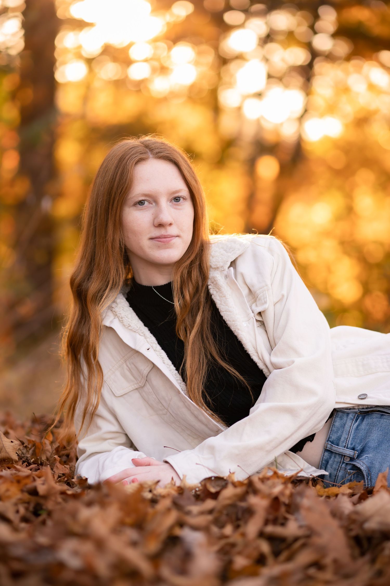 a young woman with long red hair in a white jacket lies on her elbow amid fallen autumn leaves, with beautiful autumn scenery in background