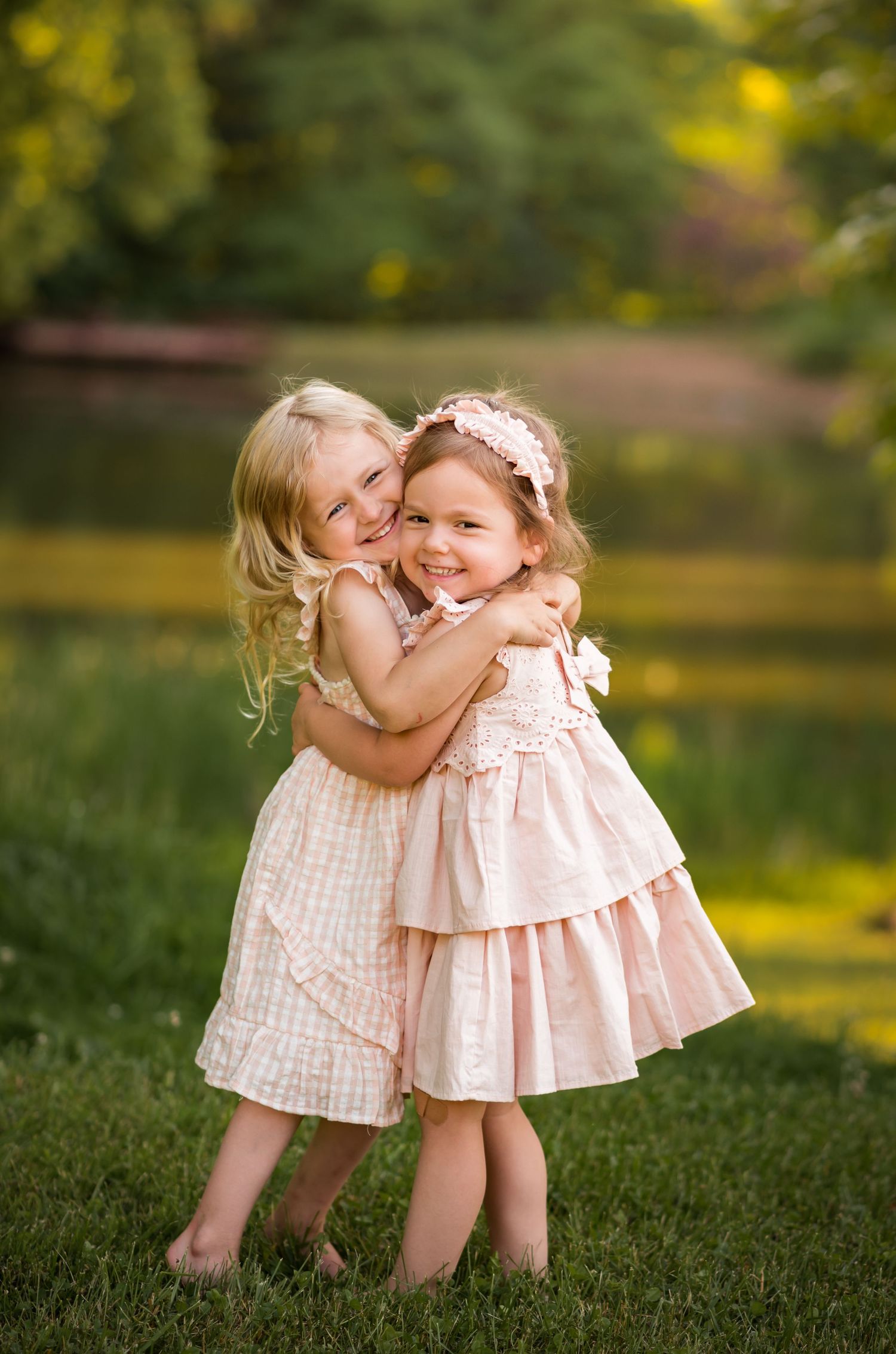 two little girl friends in pink dresses hug and giggle in a field of grass, with trees and a pond behind them
