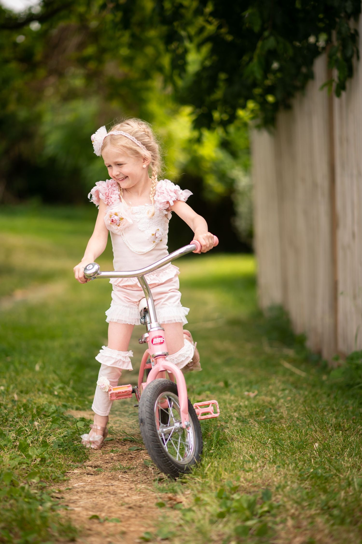 a little blonde girl in a frilly pink outfit rides a pink bicycle through the grass and laughs