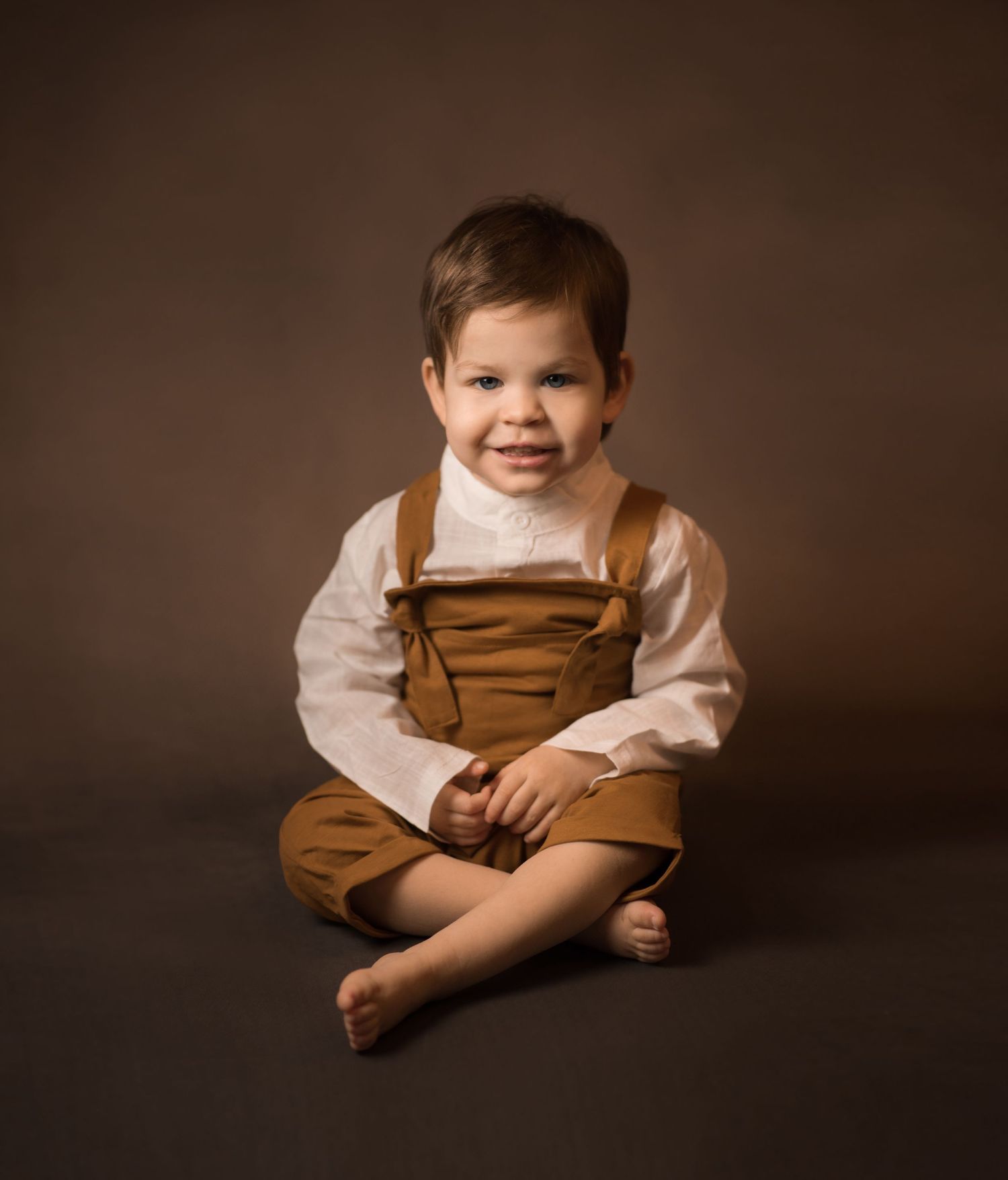 classical portrait of a little toddler boy in brown overalls, posing seated and smiling against a brown old master's studio backdrop