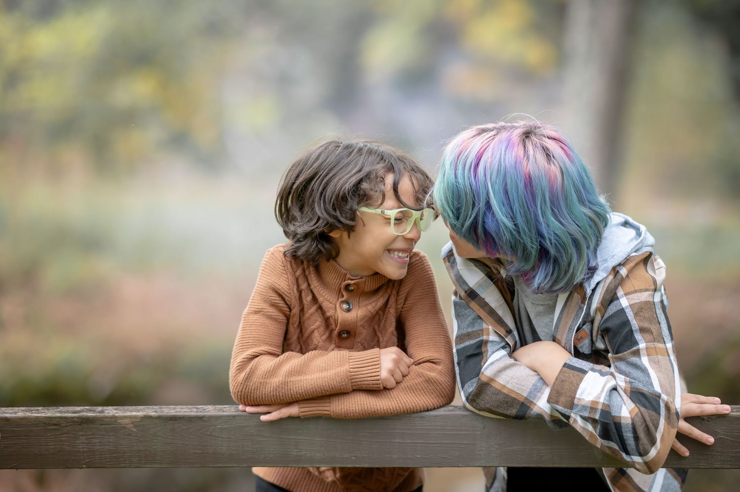 Two brothers, one with colorful glasses and one with colorful hair, share a joyful moment leaning on a wooden fence outdoors.