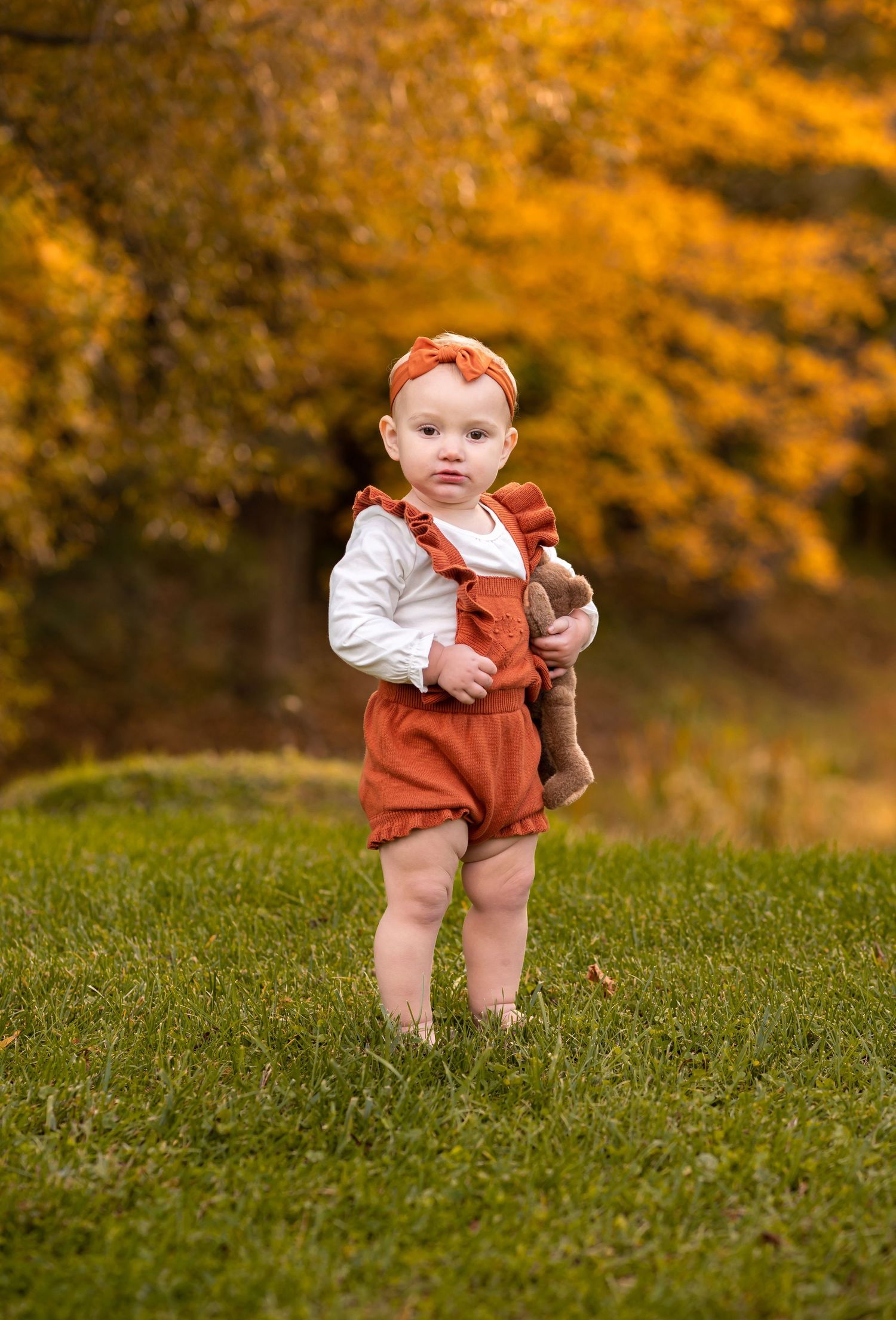 a little toddler girl in a white top and orange overalls stands in front of fall scenery in the grass holding a brown teddy bear