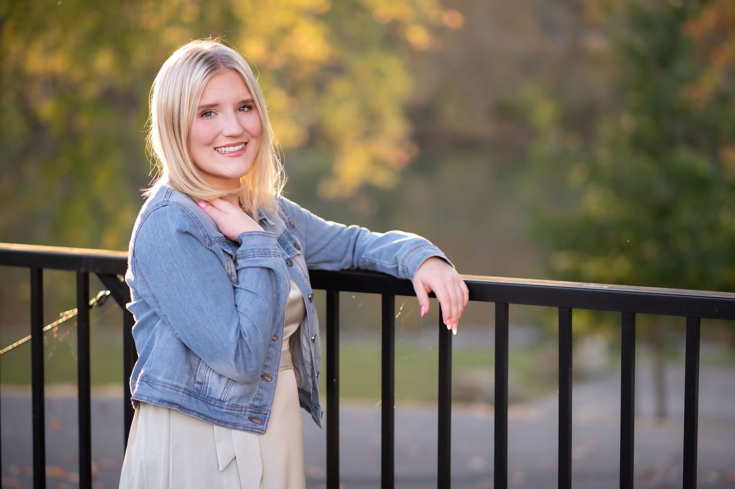 portrait of a blonde high school student in a denim jacket, leaning one arm on a railing by the river and smiling at the camera