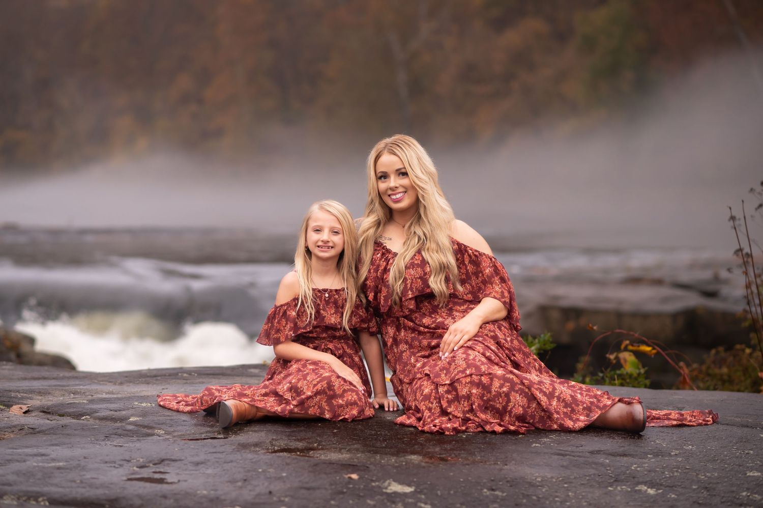 a lovely blonde mother and daughter in matching red floral dresses pose seated on a large rock cliff with fog and a foaming waterfall behind