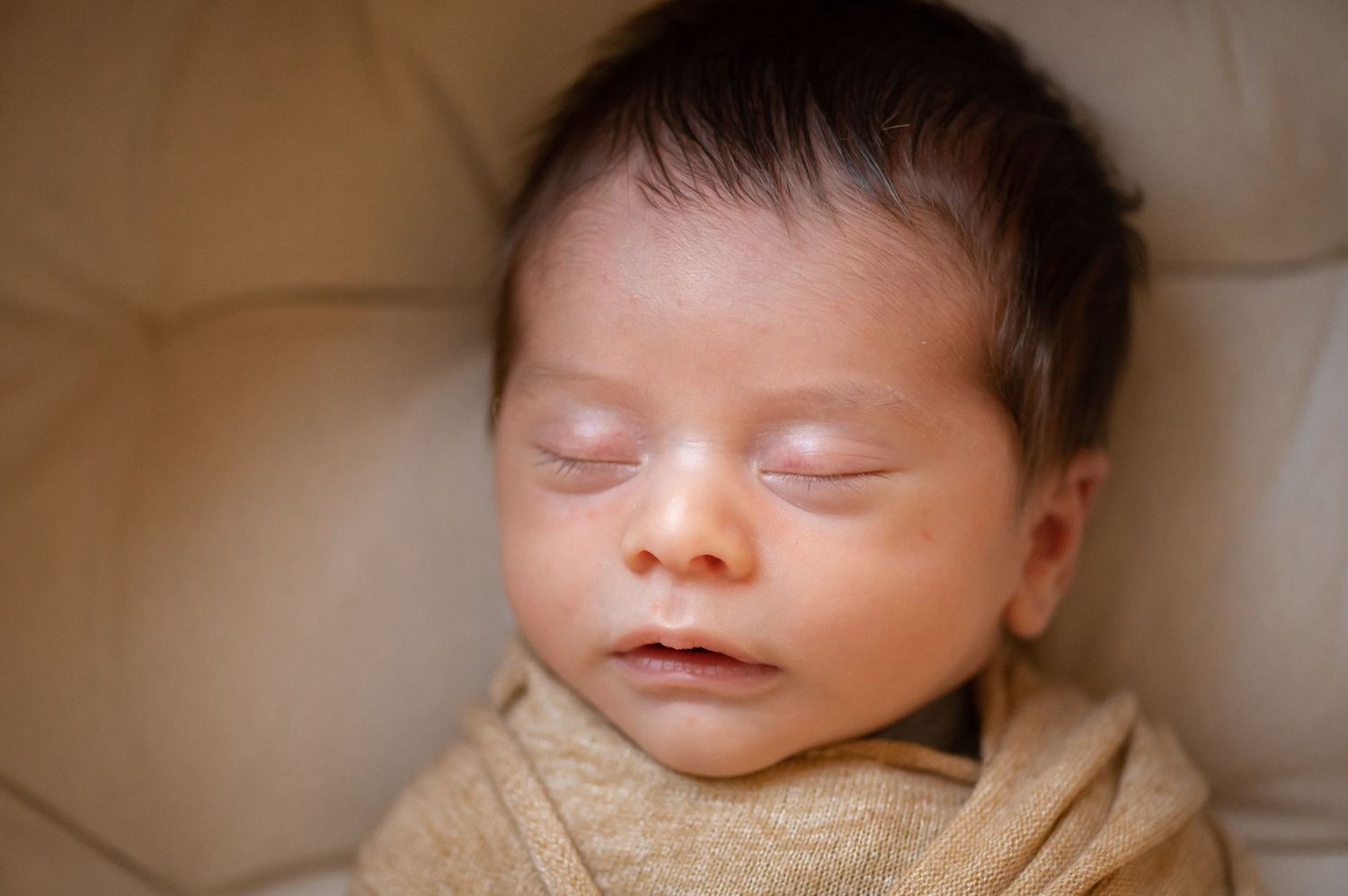 Peaceful newborn baby sleeping swaddled in a warm golden wrap, with dark hair and serene expression.