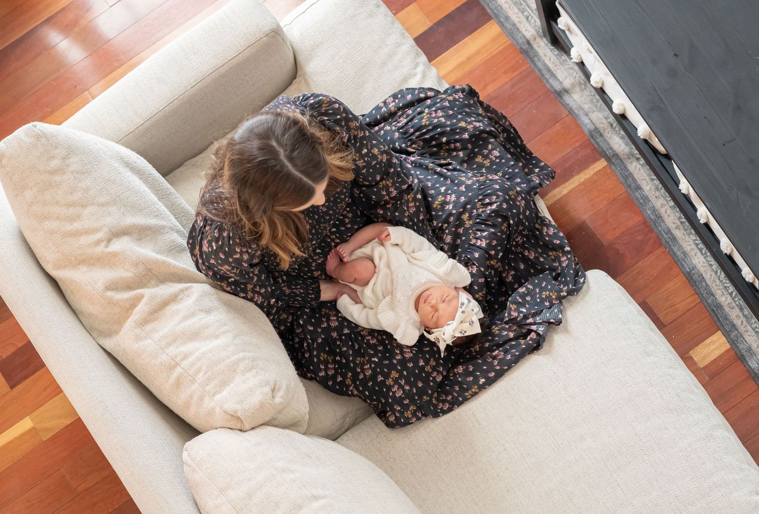 Aerial view of a mother in floral dress cradling her newborn baby on a cream sofa indoors.