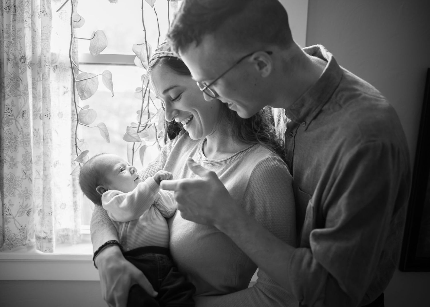 black and white portrait of a young couple standing together in front of a window, holding and smiling at their newborn baby.
