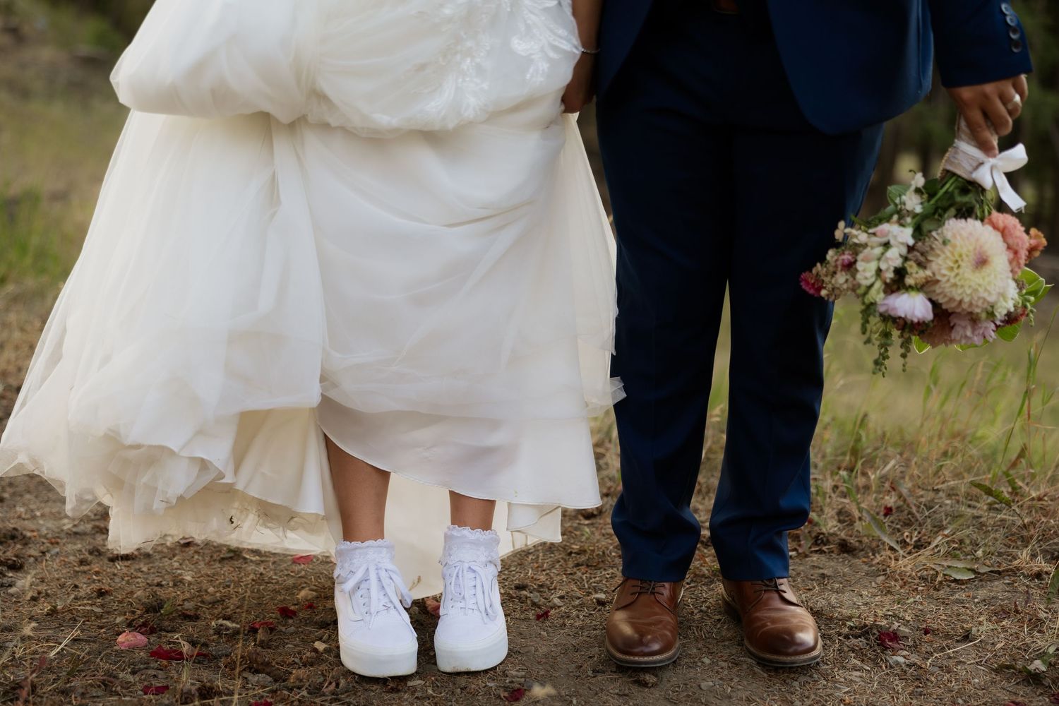 Wedding couple's feet showing white sneakers and brown dress shoes on grass.